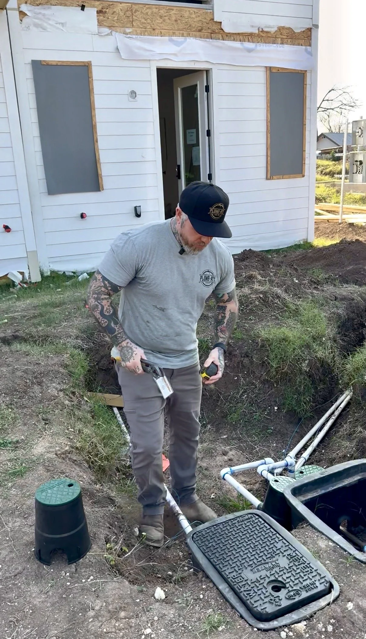 A man with tattoos and a baseball cap working on underground plumbing outside a house, inspecting the pipes. Construction materials and tools are visible around him.