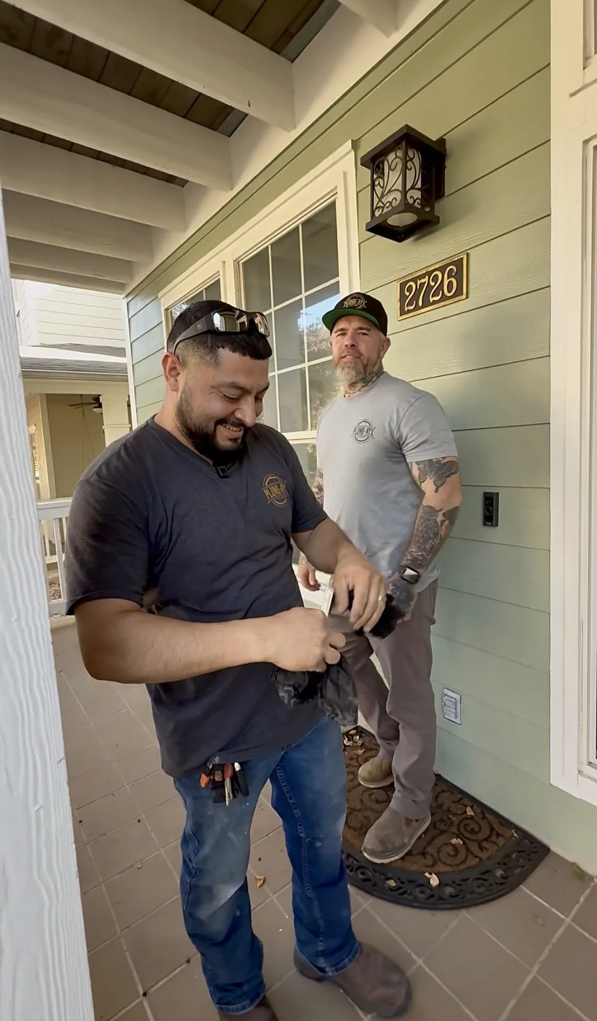 Two men standing on a porch outside a house, one looking down and smiling while the other looks at camera, with green siding, a window, a wall-mounted light, house number 2726, and a decorative doormat behind them.