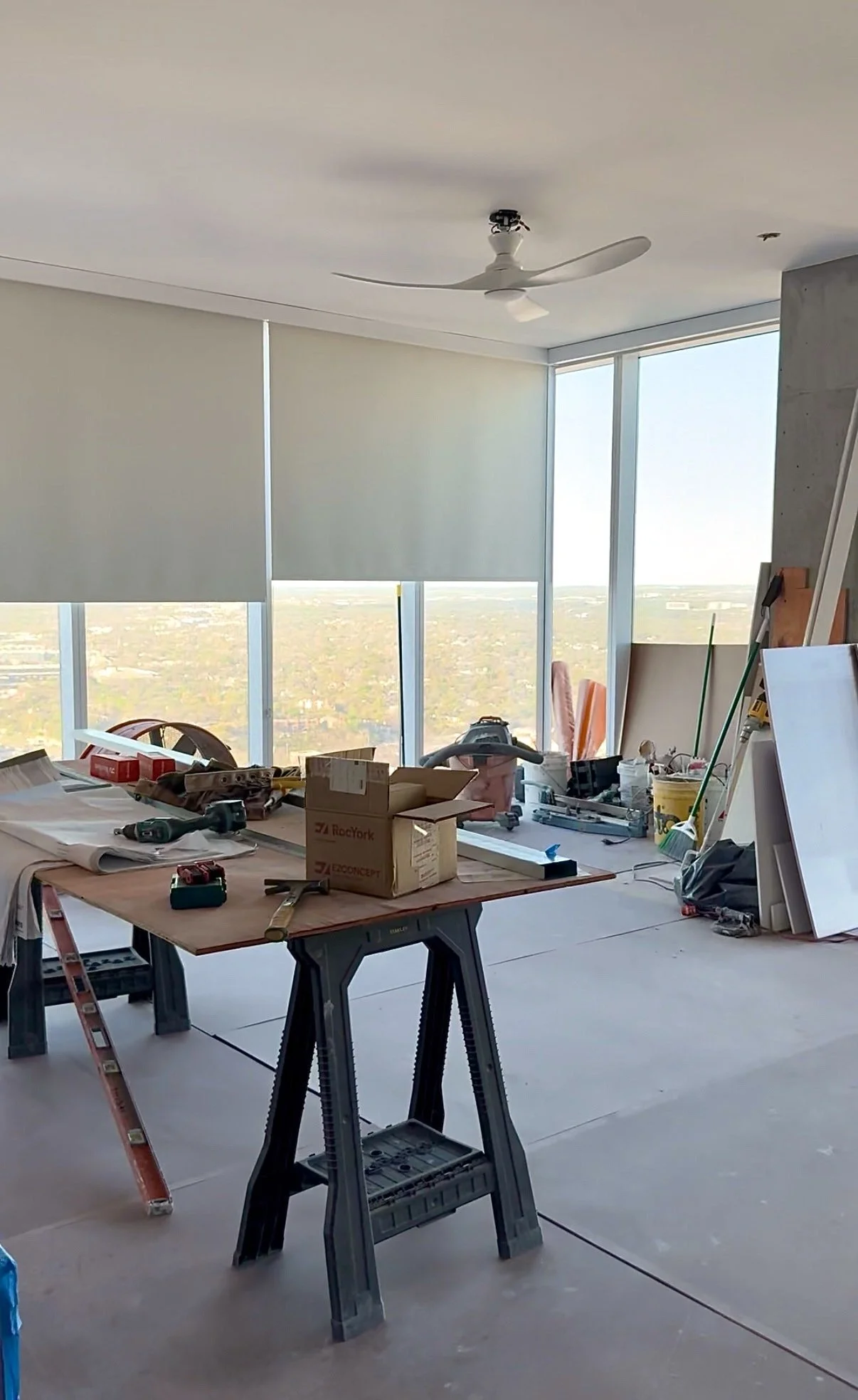 Interior of a high-rise building under construction with tools, construction materials, and a work table, large windows showing a city view, ceiling fan without blades.
