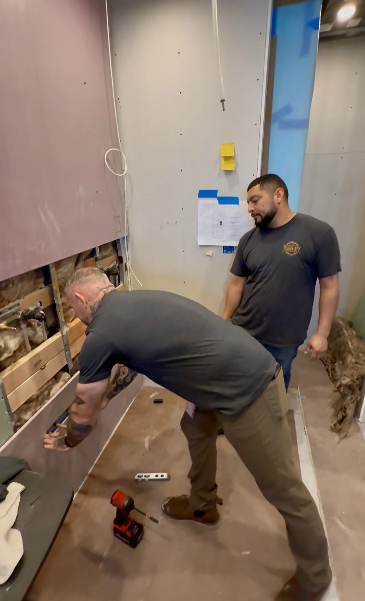 Two men working on a plumbing project in a construction area, installing or repairing pipes inside a wall. One man is bending down to fit pipes, while the other watches.
