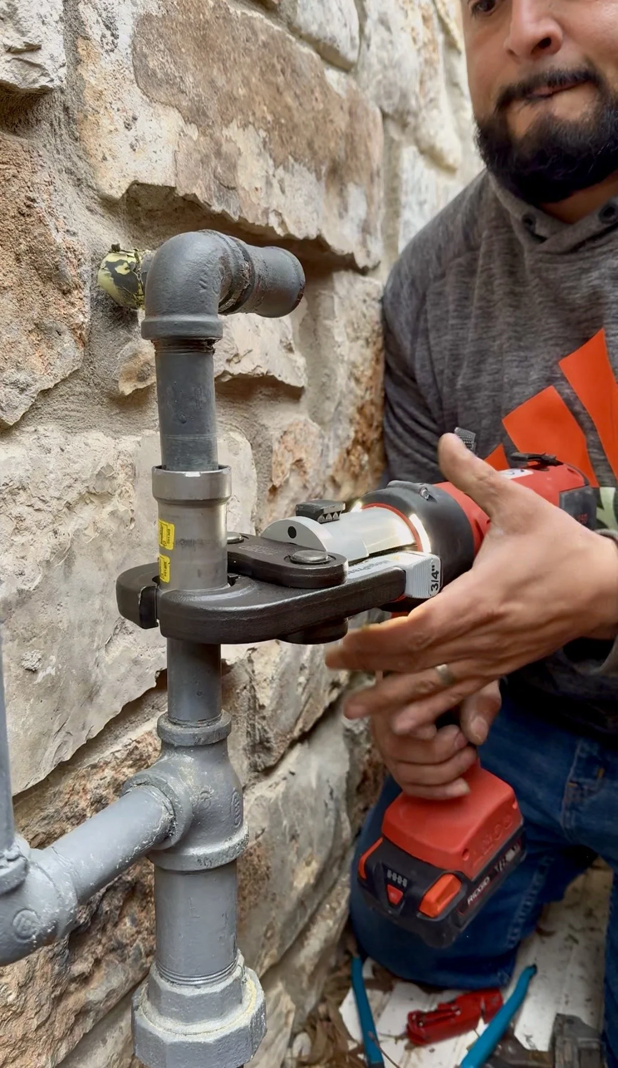 A person using a cordless power drill to work on plumbing pipes mounted on a brick wall.