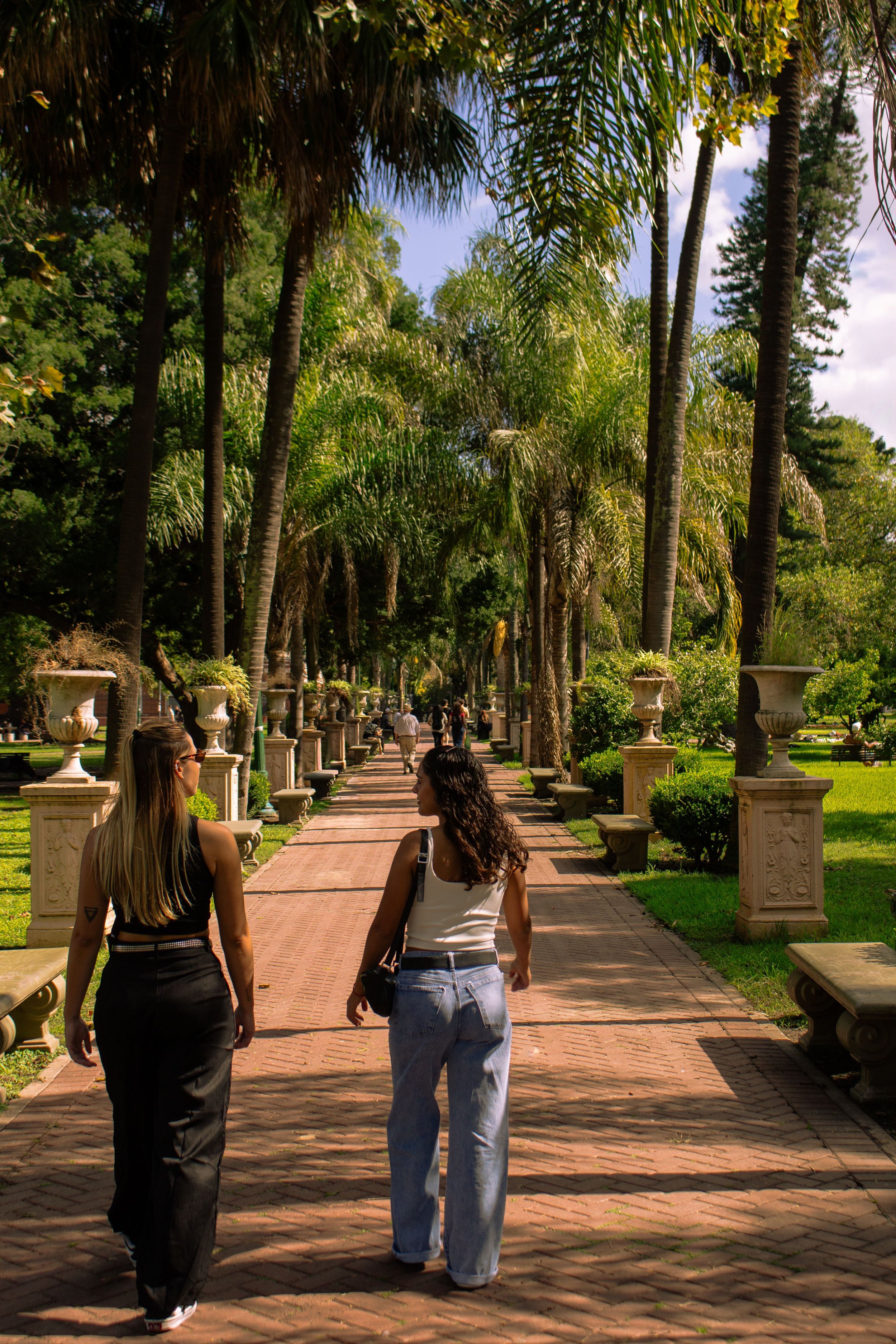 Two young women walking and talking in a park with palm trees, stone benches, and decorative planters. The sky is partly cloudy and sunny.