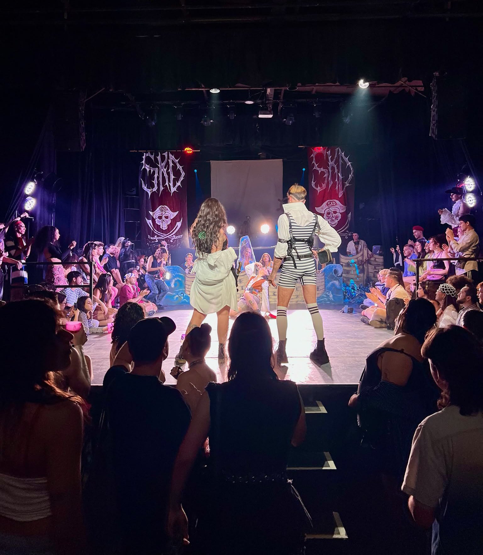 A large group of people gathered in an indoor theater or performance space, watching a live stage performance with a band or musical group. The stage has bright lights, banners with skulls, and two central performers facing the audience.