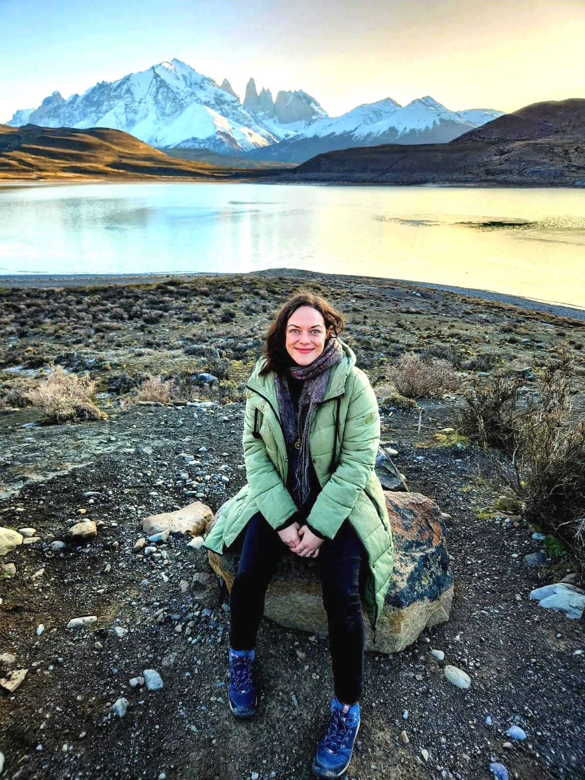 A woman sitting on a rock near a lake with snow-capped mountains in the background, wearing a green jacket and smiling.