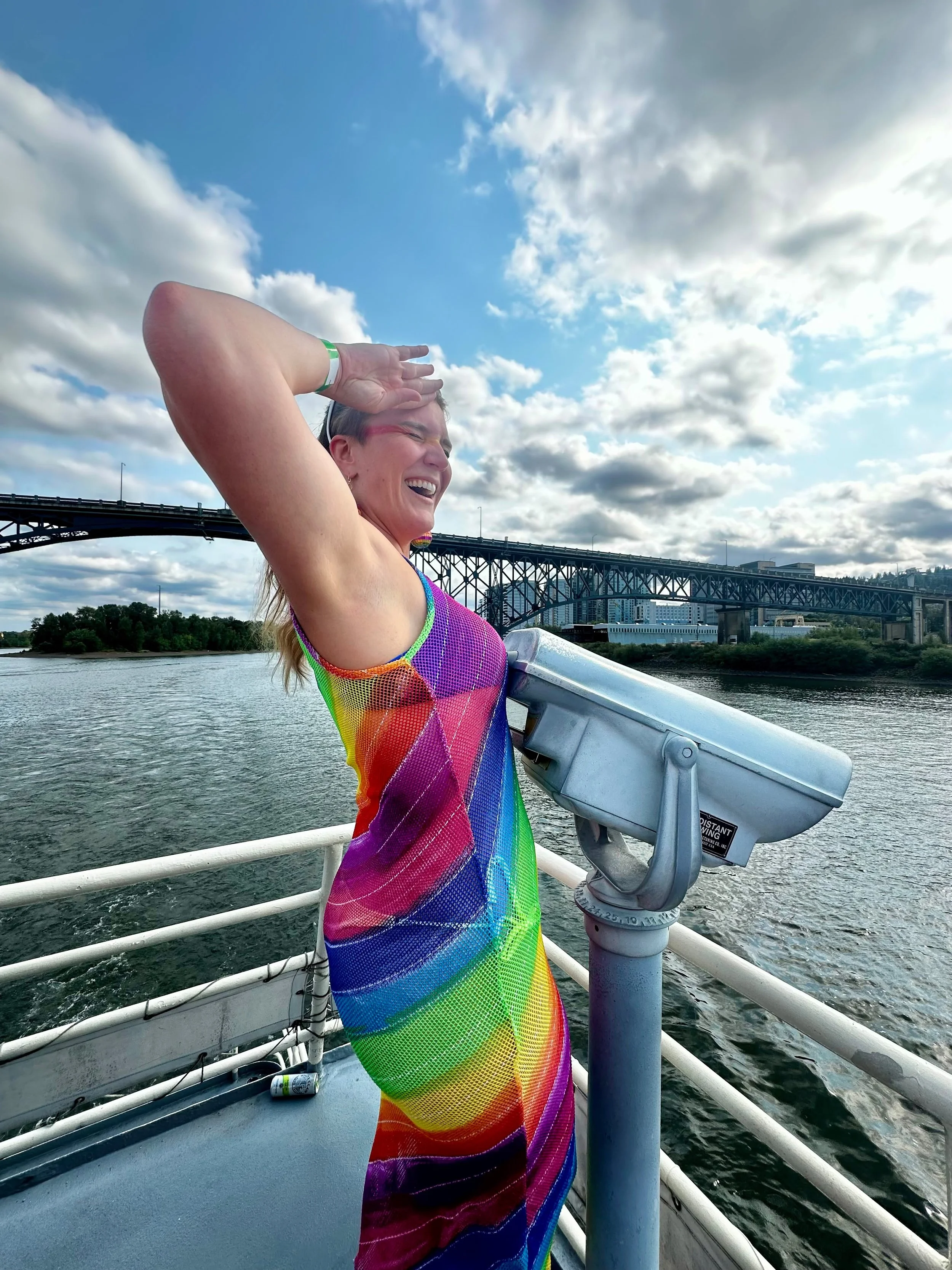 Woman wearing a rainbow-striped dress on a boat, looking out over the water with a bridge and city skyline in the background, and sky with scattered clouds.