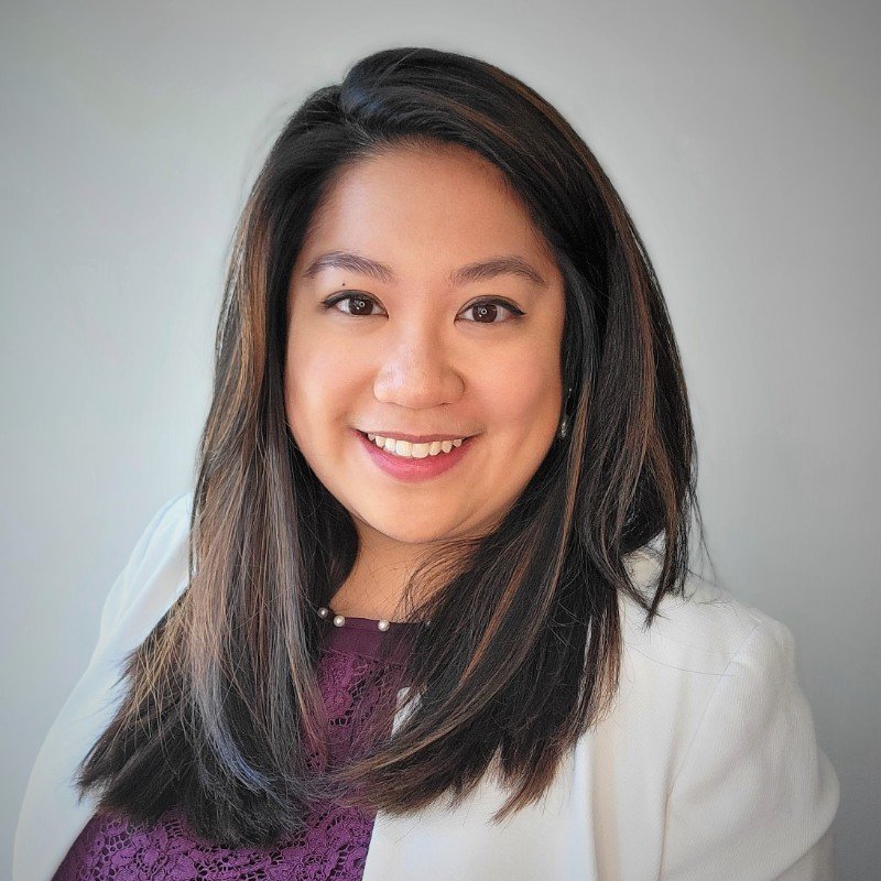 Portrait of a smiling woman with long dark hair, wearing a white blazer and a purple top, against a neutral background.