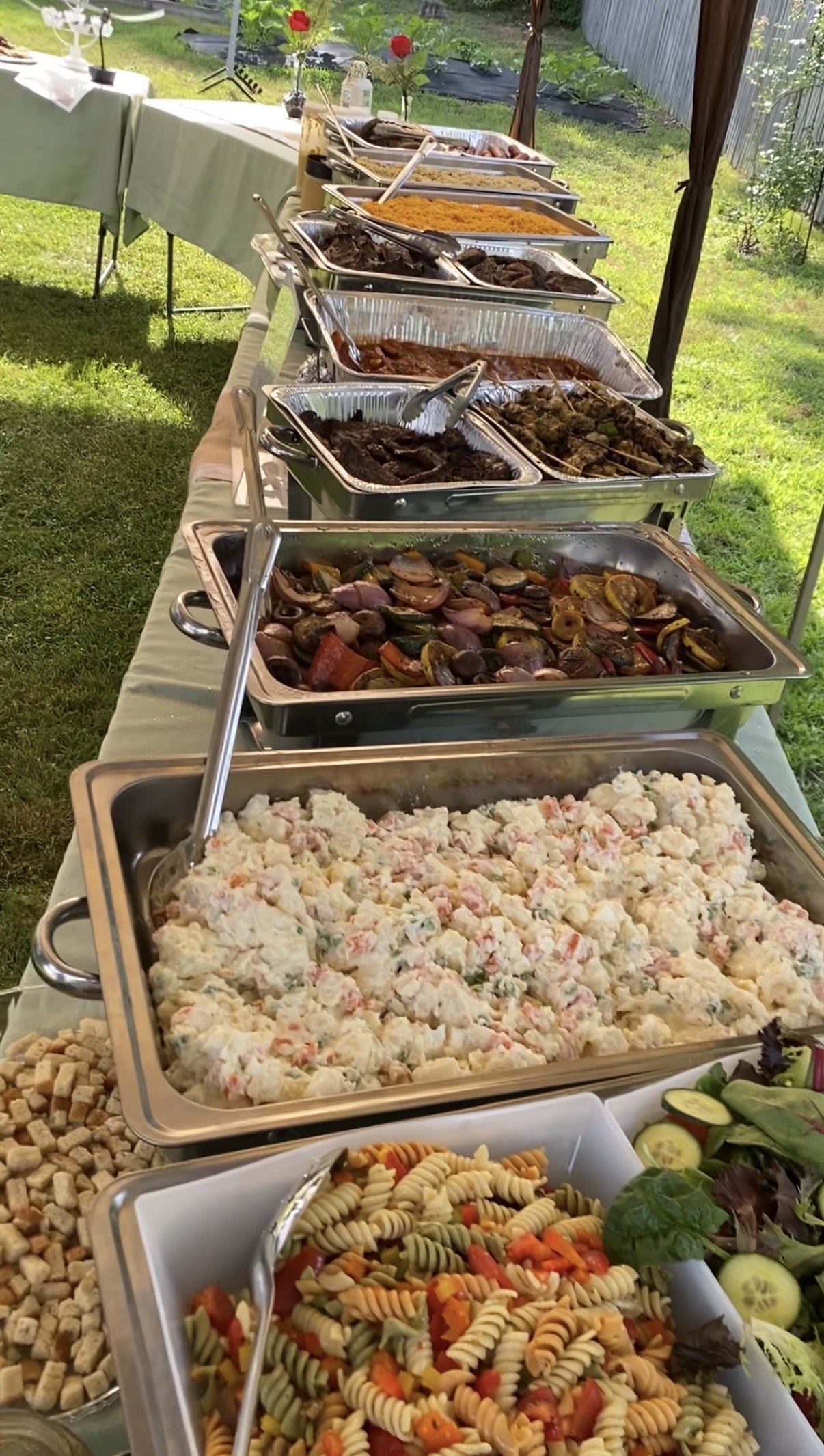 Buffet table with various salads, pasta, and cooked dishes in aluminum trays outdoors on a green lawn.