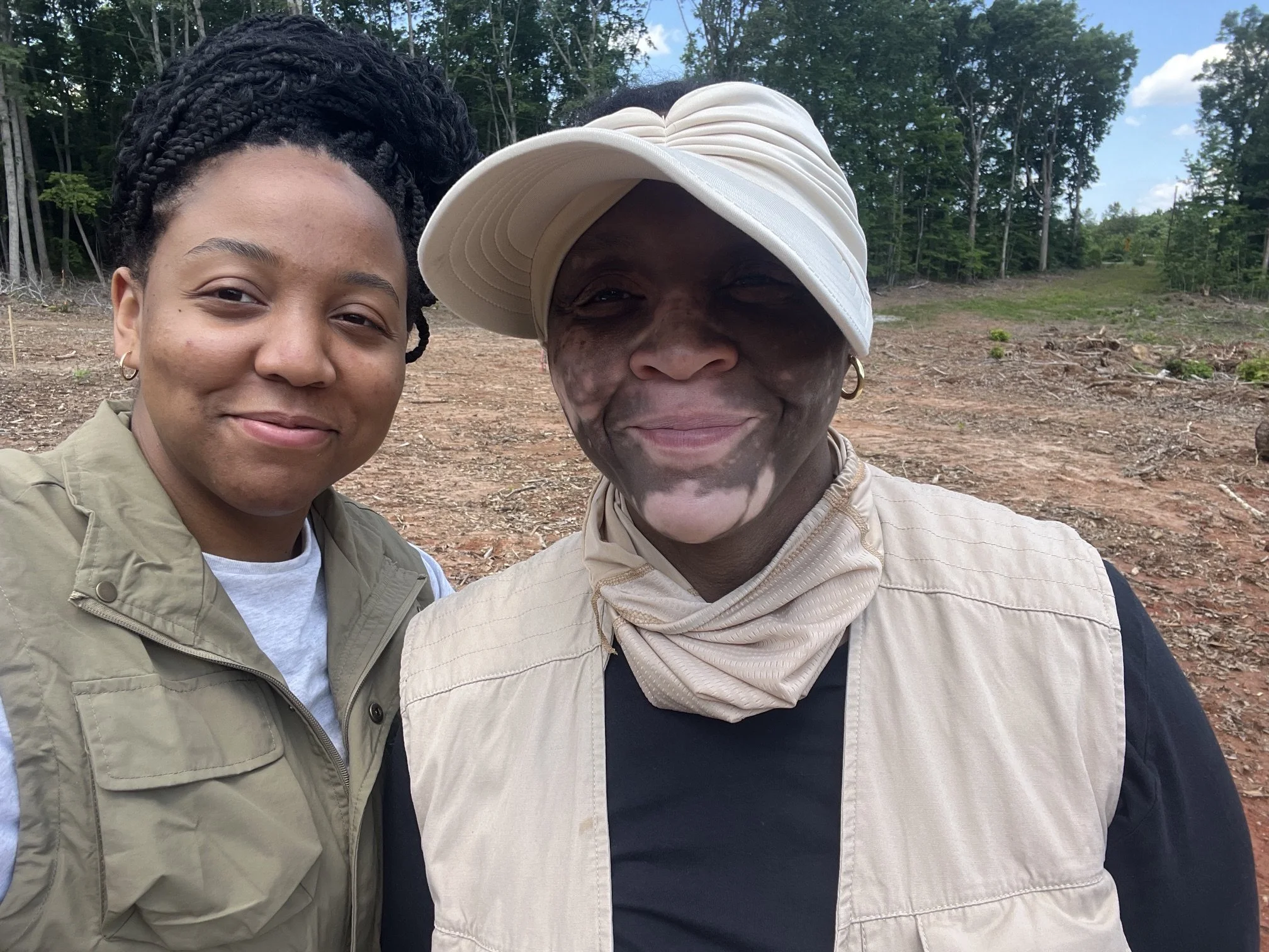 Two women are standing outdoors in a natural setting with trees and a dirt ground visible behind them. They are close together and smiling at the camera. One woman has dark hair styled in braids, is wearing a tan jacket, and has hoop earrings. The other woman has dark skin, is wearing a large white hat tilted to one side, a scarf around her neck, and earrings.