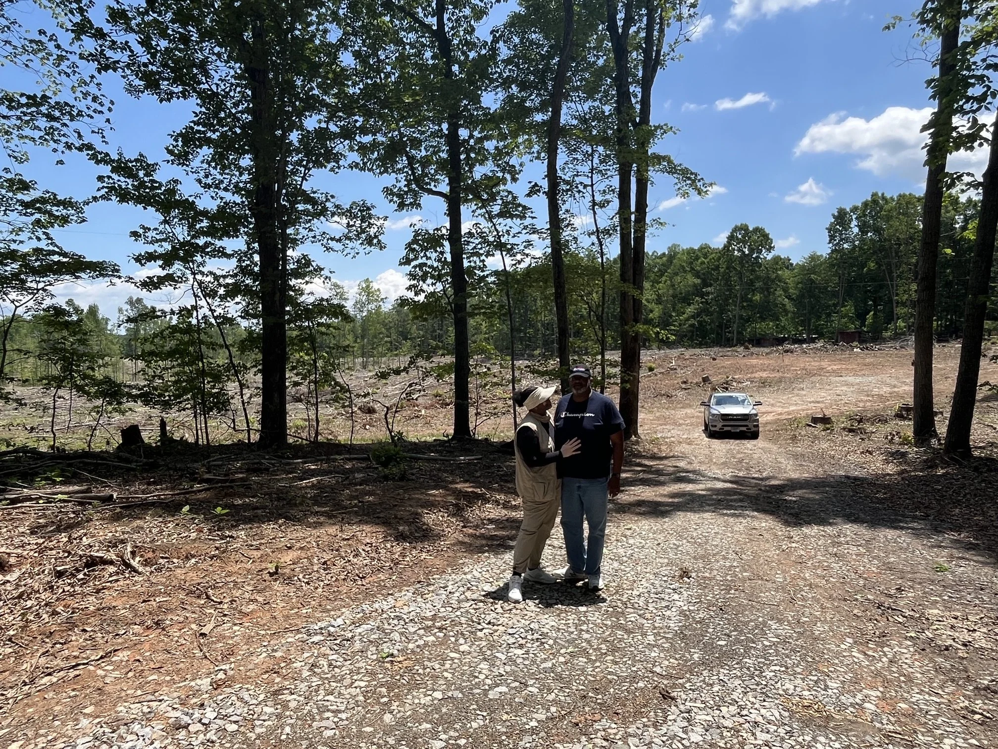 Two people standing outdoors in a cleared, wooded area during daytime, with a dog and a pickup truck nearby.