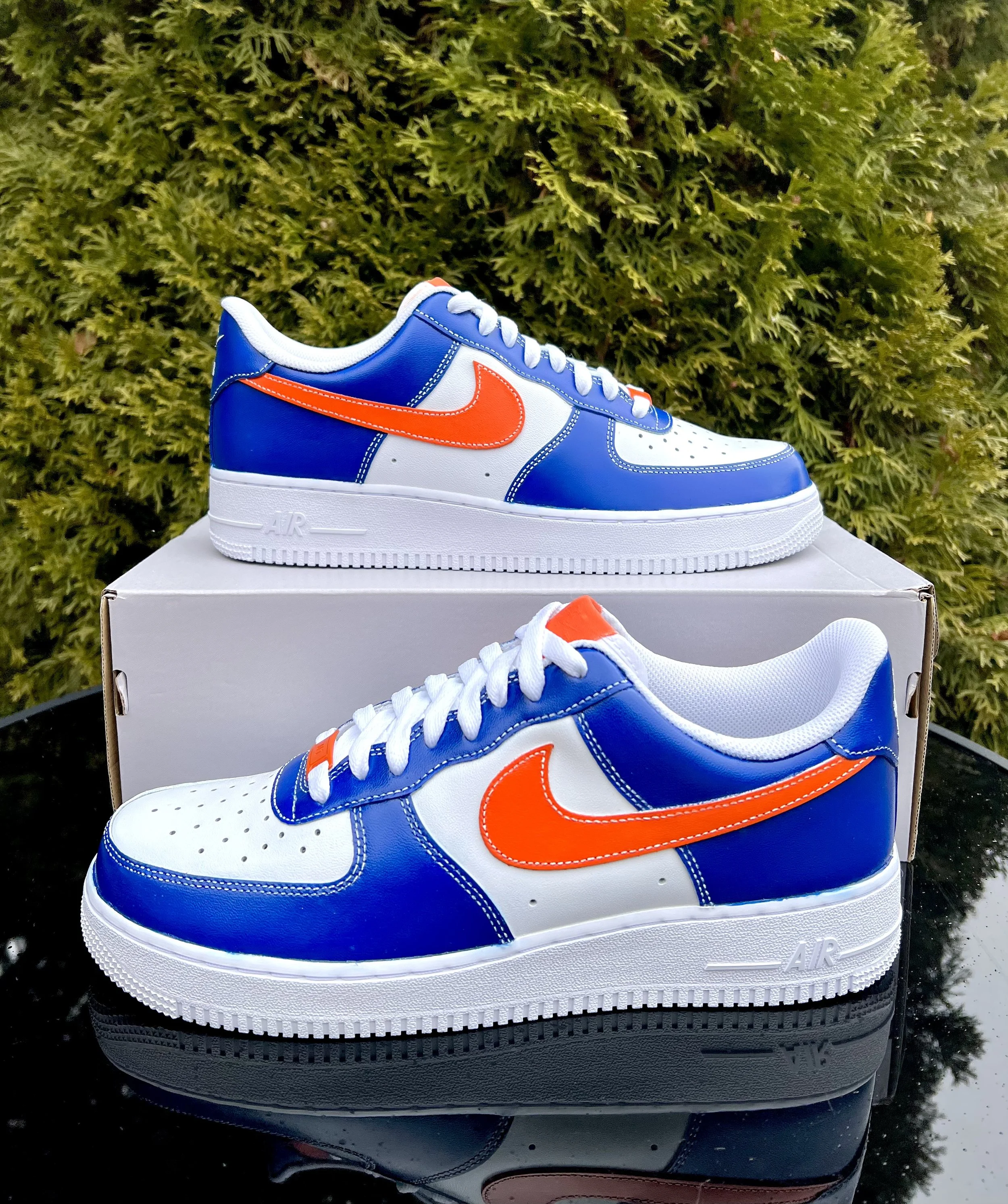 Blue and white Nike AirForce 1 sneaker with orange Nike swoosh, placed on a cardboard shoe box and reflected on a black glossy surface with greenery background.