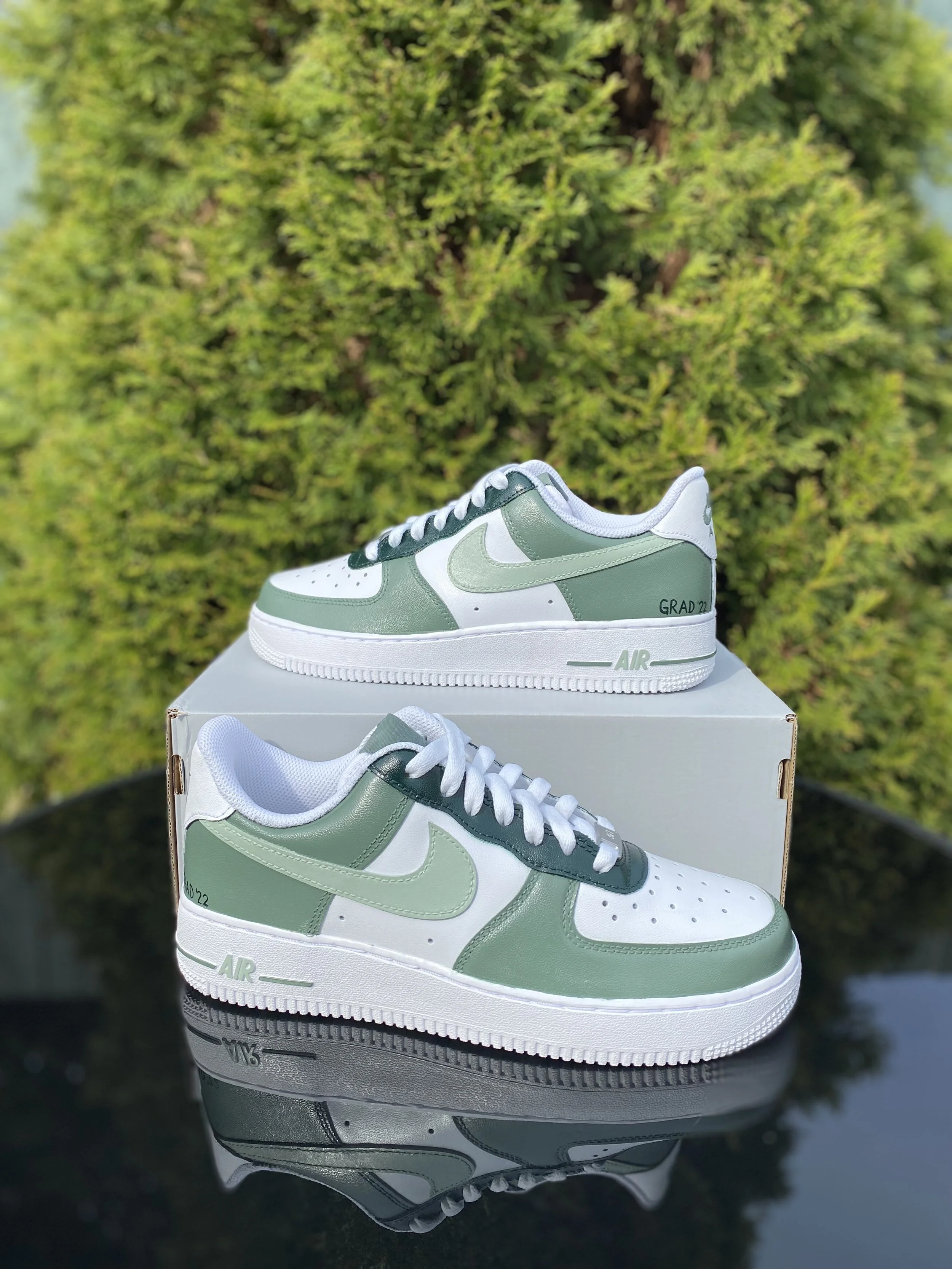 A pair of green and white Nike sneakers on a white shoebox, reflected on a glossy black surface, with a background of green bushes and trees.