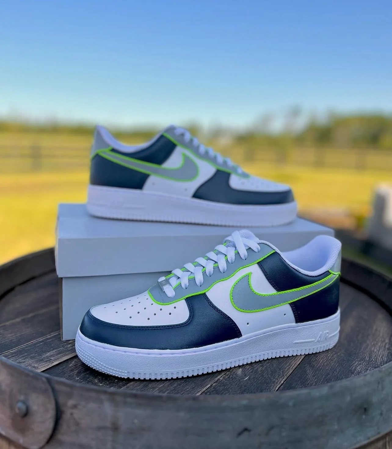 A pair of Nike Air Force 1 sneakers with white, navy, and neon green accents, positioned on a circular wooden surface with a blurred outdoor background.