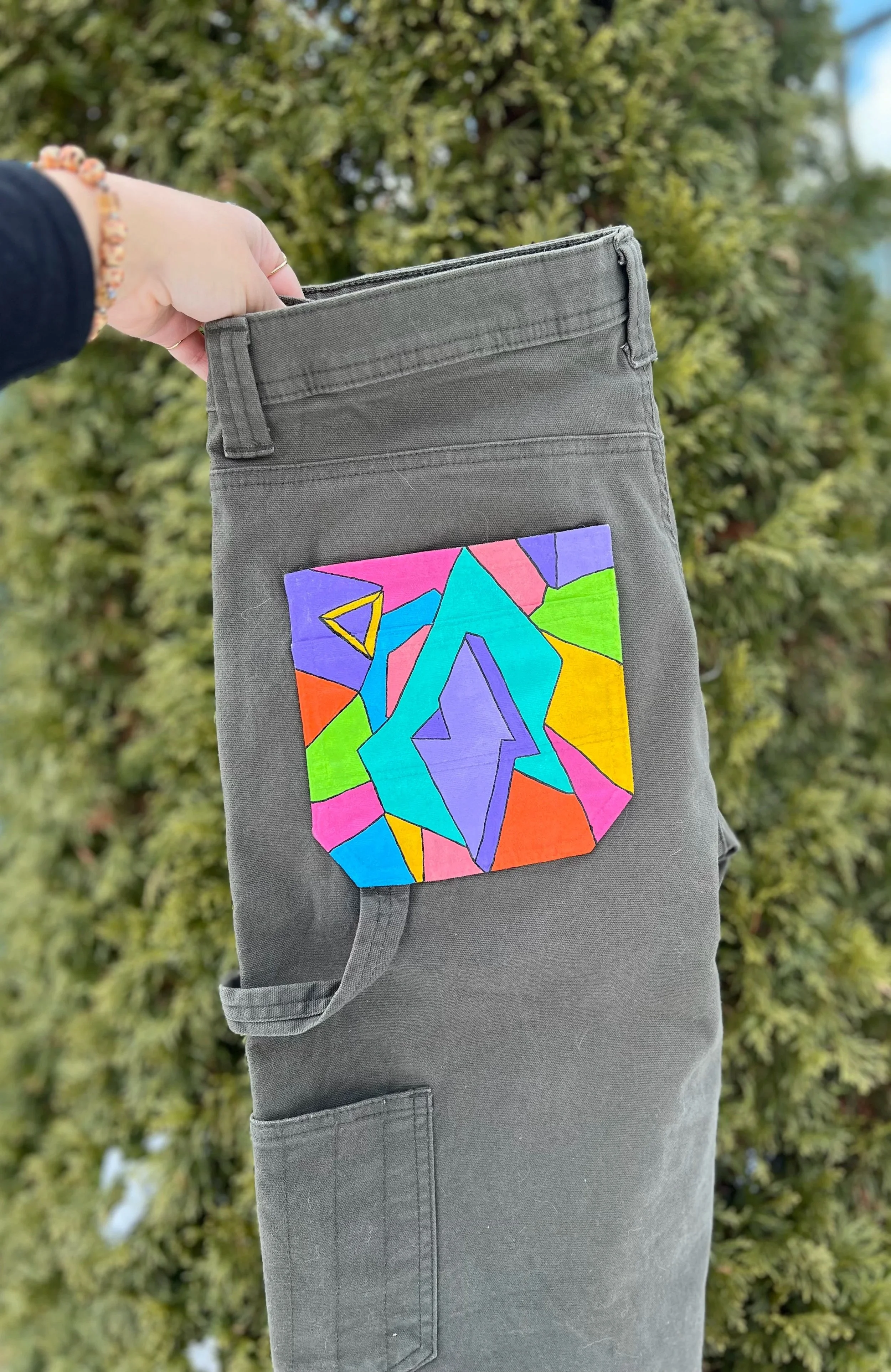 A person holding a gray cargo pant leg with a colorful geometric design patch pocket, standing outdoors with trees in the background.