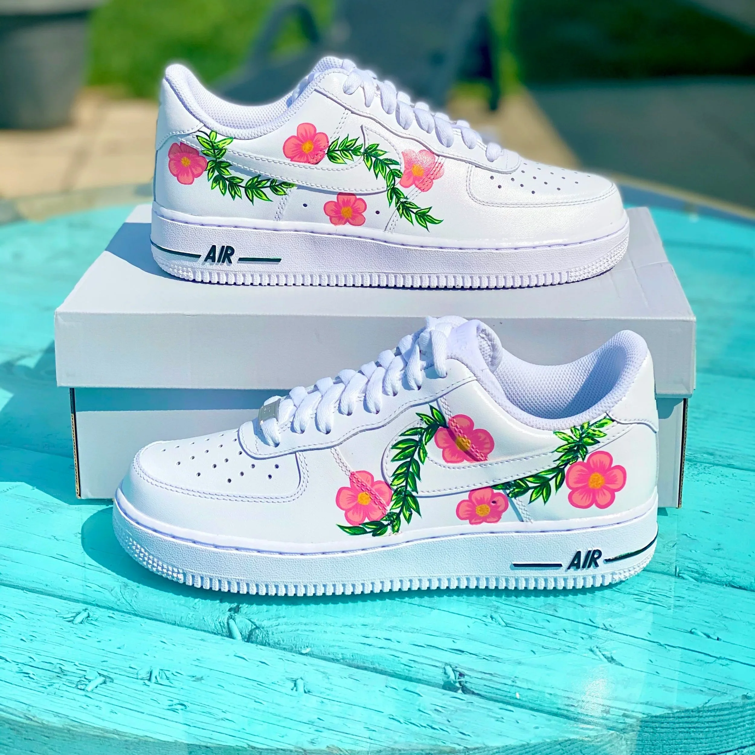 White Nike Air Force 1 sneakers with pink floral and green leaf designs, placed on a white shoebox on a turquoise wooden surface.
