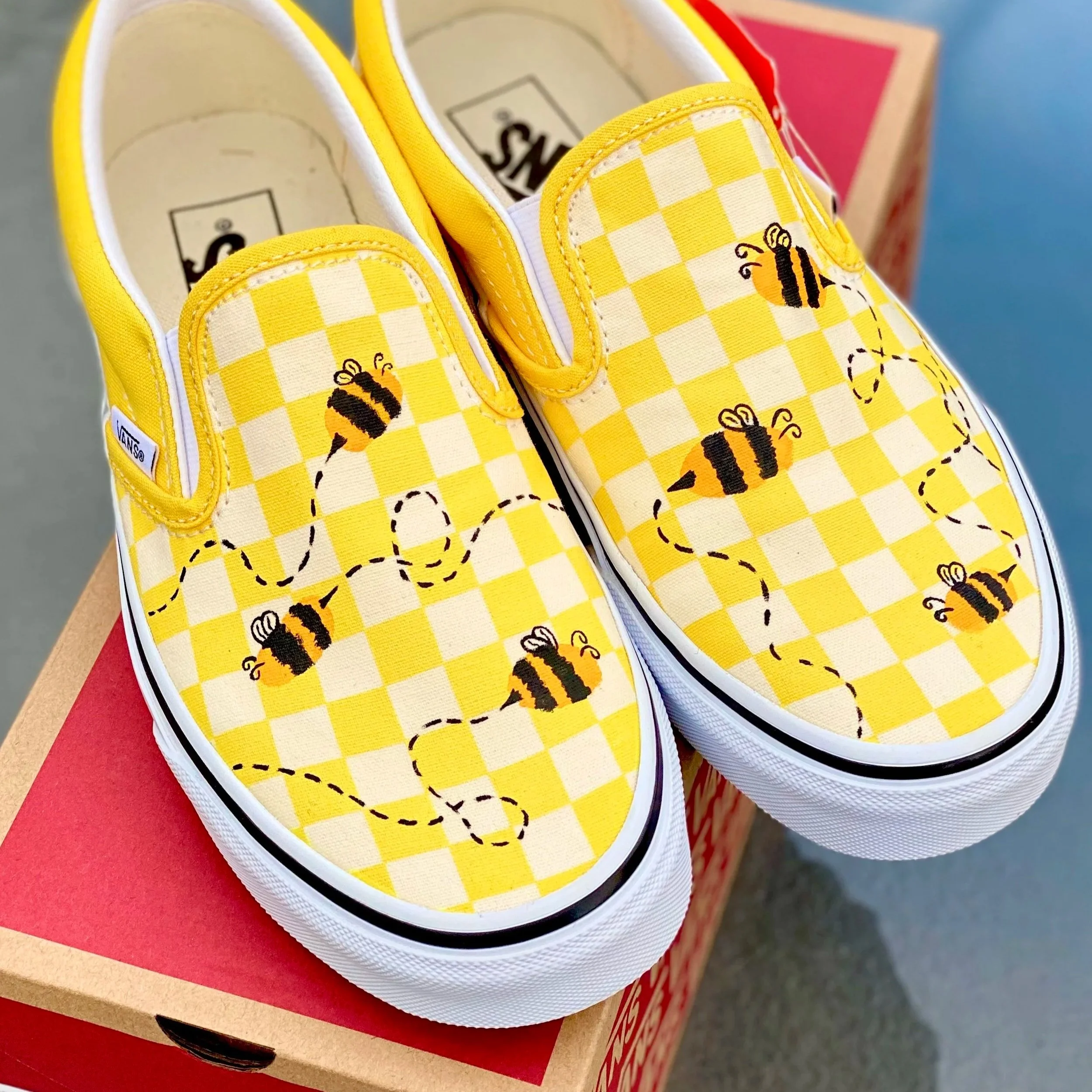 A pair of yellow Vans slip-on shoes with a checkered pattern and embroidered bees on the top, placed on a red and brown cardboard box.