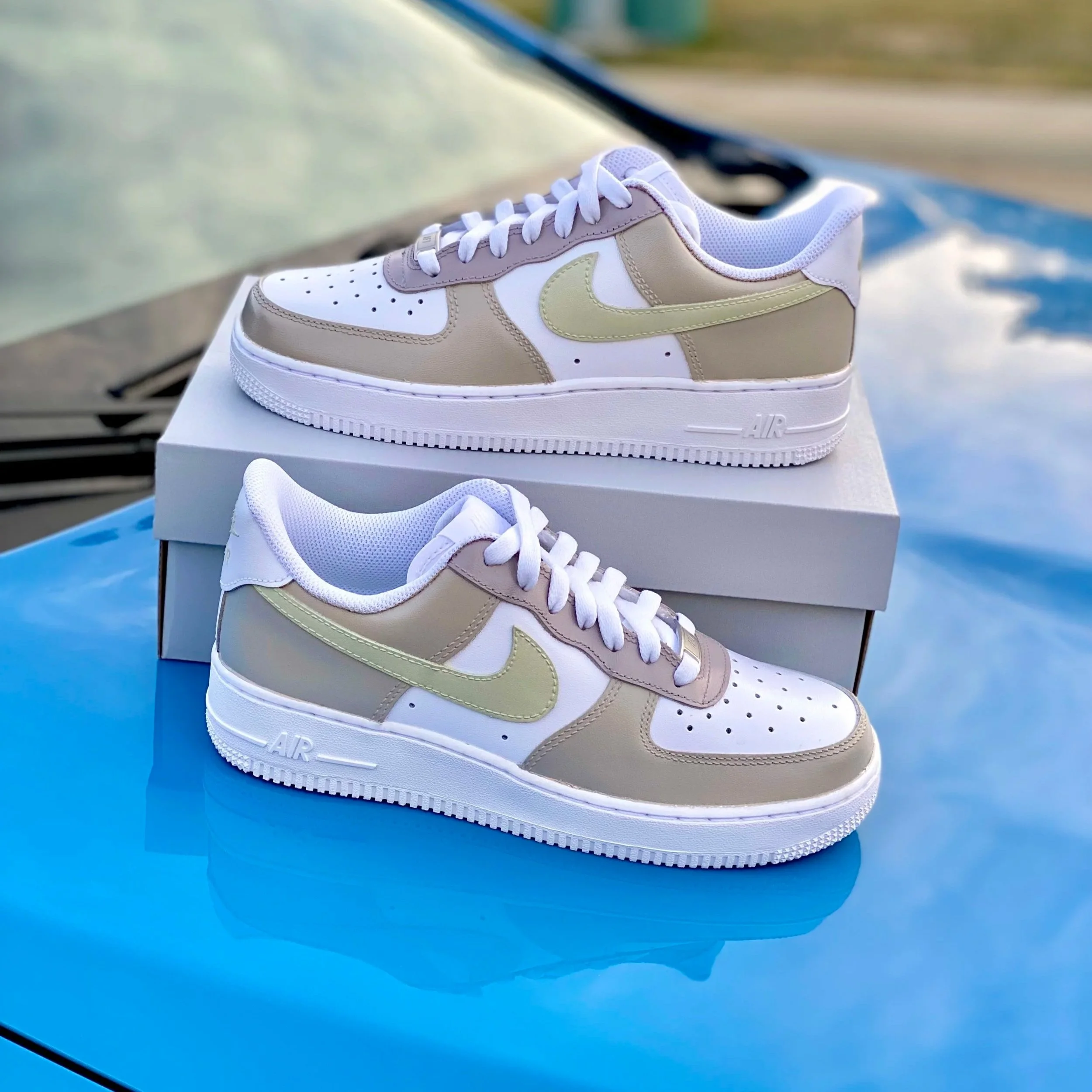 White and beige Nike Air Force 1 sneakers with white laces, placed on a white box against a blurred background.