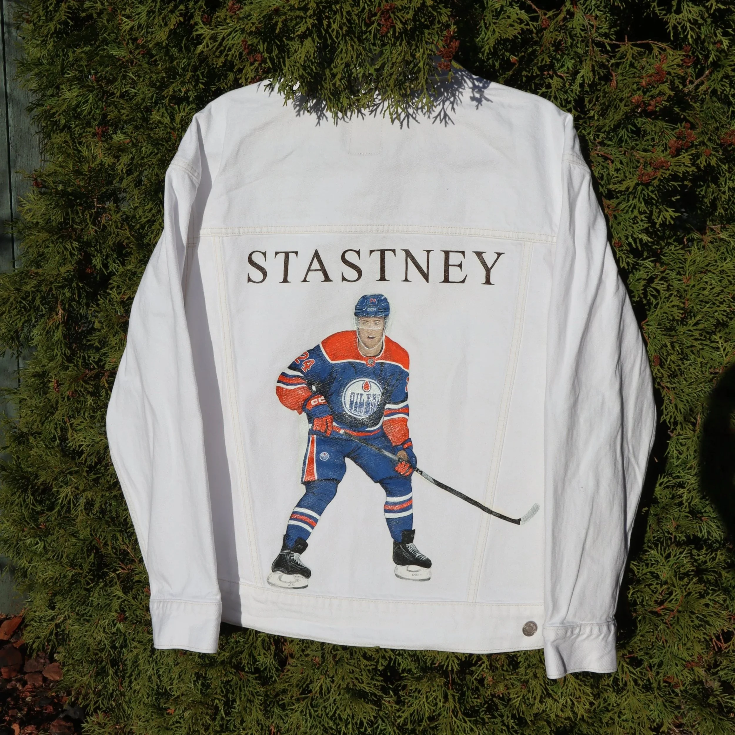White jersey with the name 'Stasney' and a cartoon image of a hockey player wearing a blue, orange, and white uniform, holding a hockey stick, standing in front of green shrubbery.