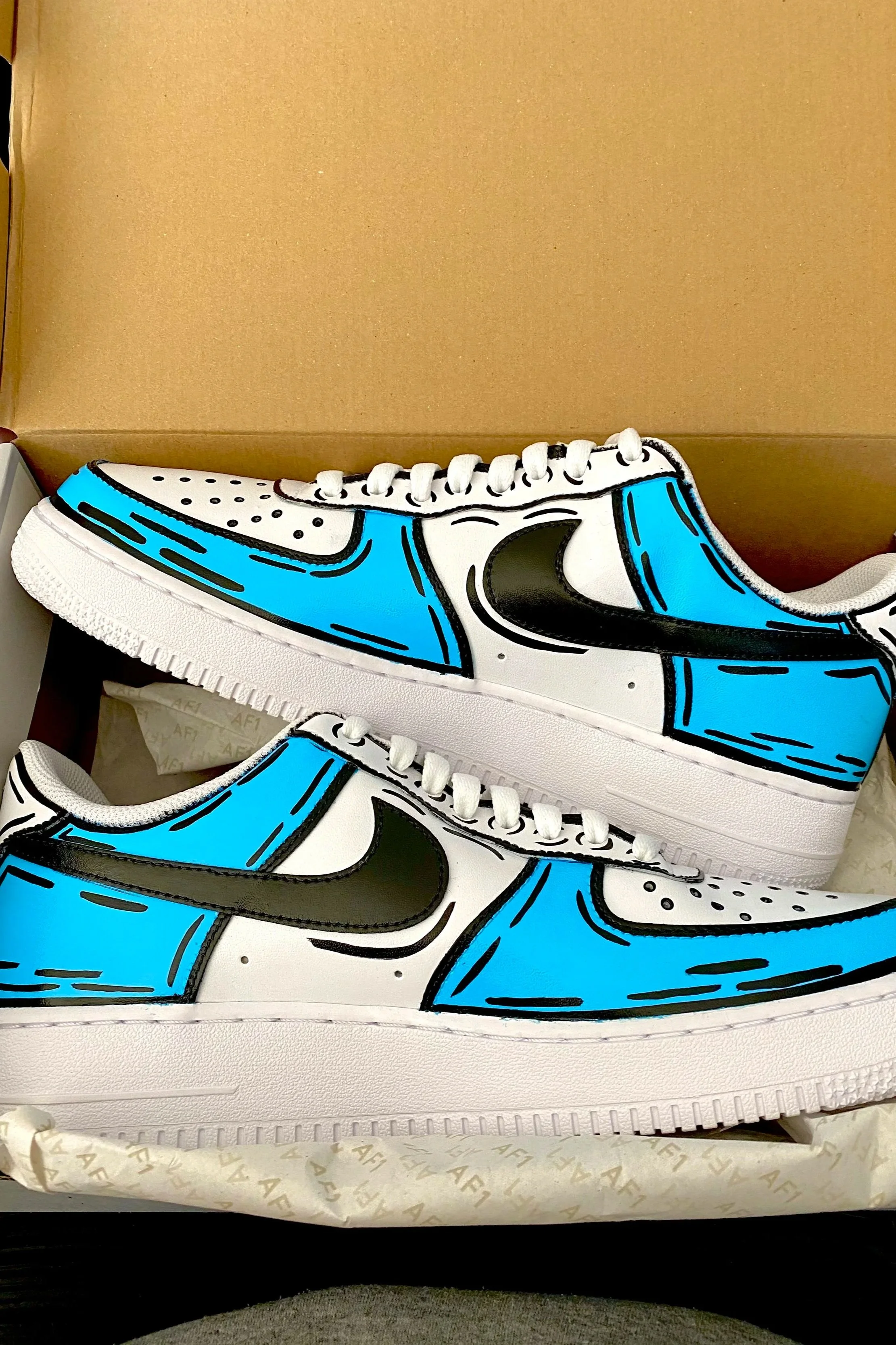 Two white sneakers decorated with black and blue outline artwork of a sneaker, inside an open shoebox with tissue paper.