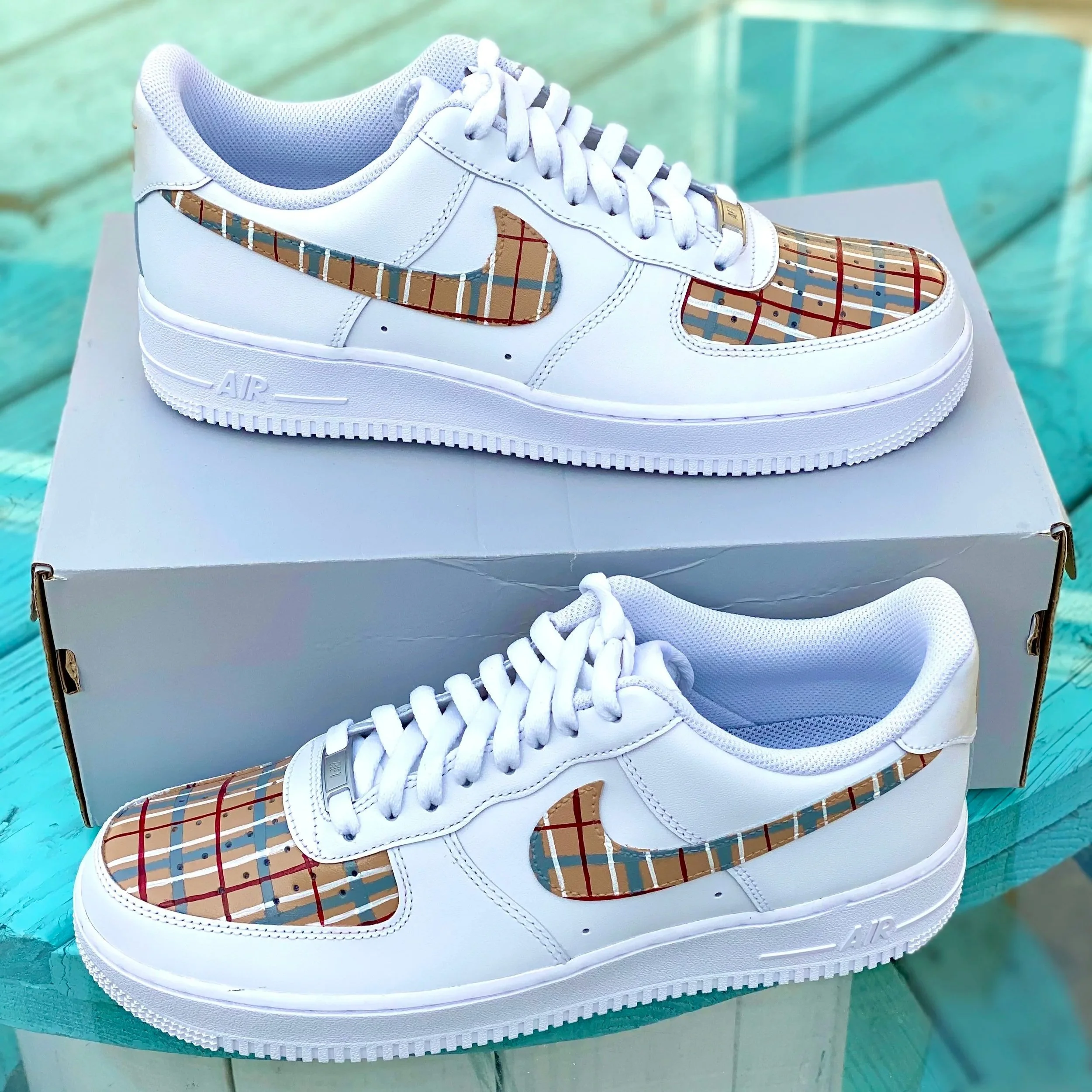 White sneaker with plaid pattern accents on the swoosh and toe area, placed on a white box outdoors against a teal wooden surface.