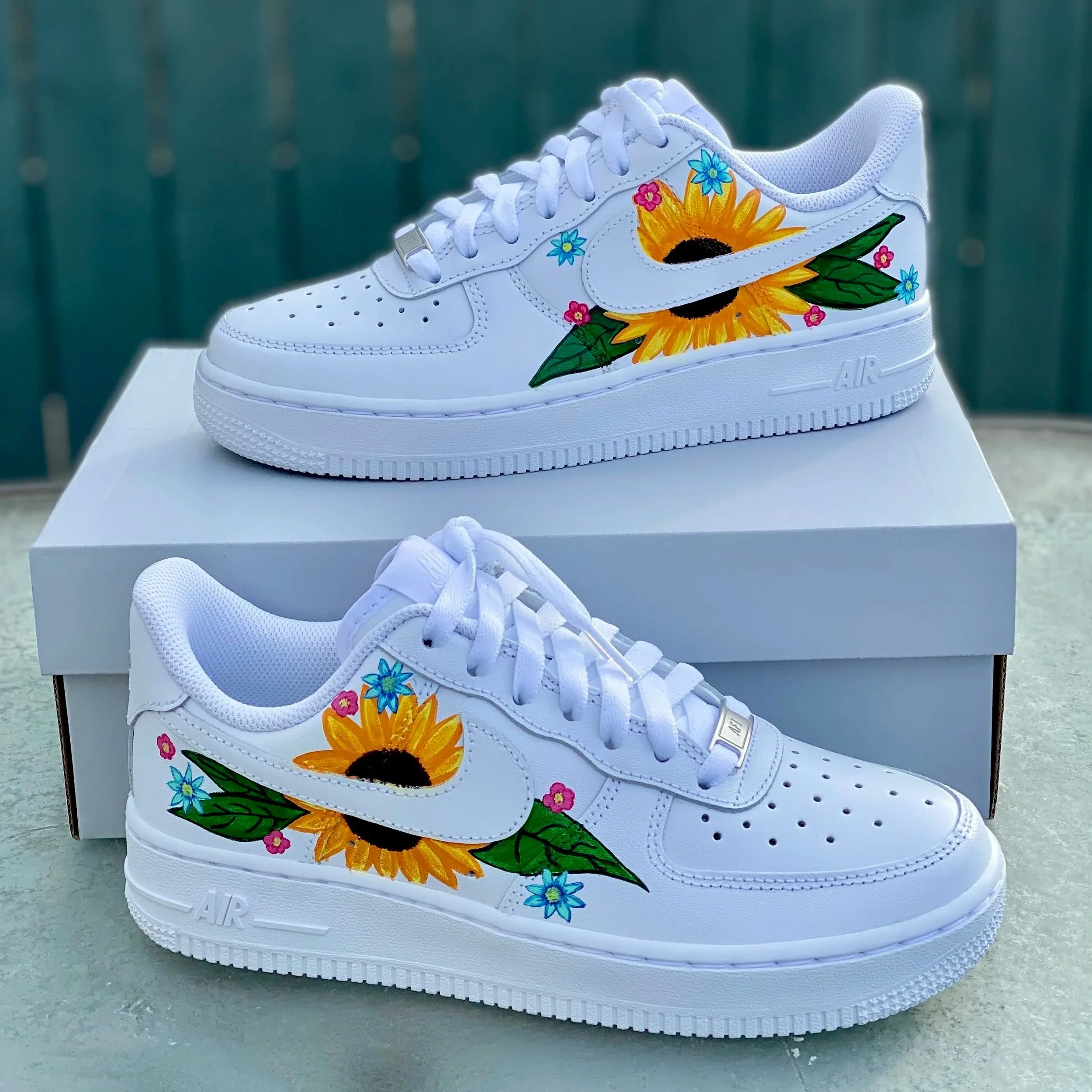 White Nike Air Force 1 sneaker with colorful hand-painted sunflower and small pink and blue flowers on the side, placed on a white box on a green outdoor surface.