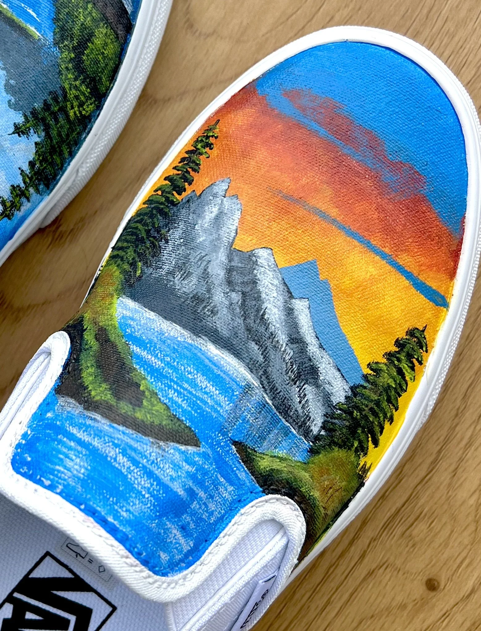 Close-up of a painted sneaker showing a landscape scene with mountains, trees, a mountain river, and a sunset sky in vibrant colors.