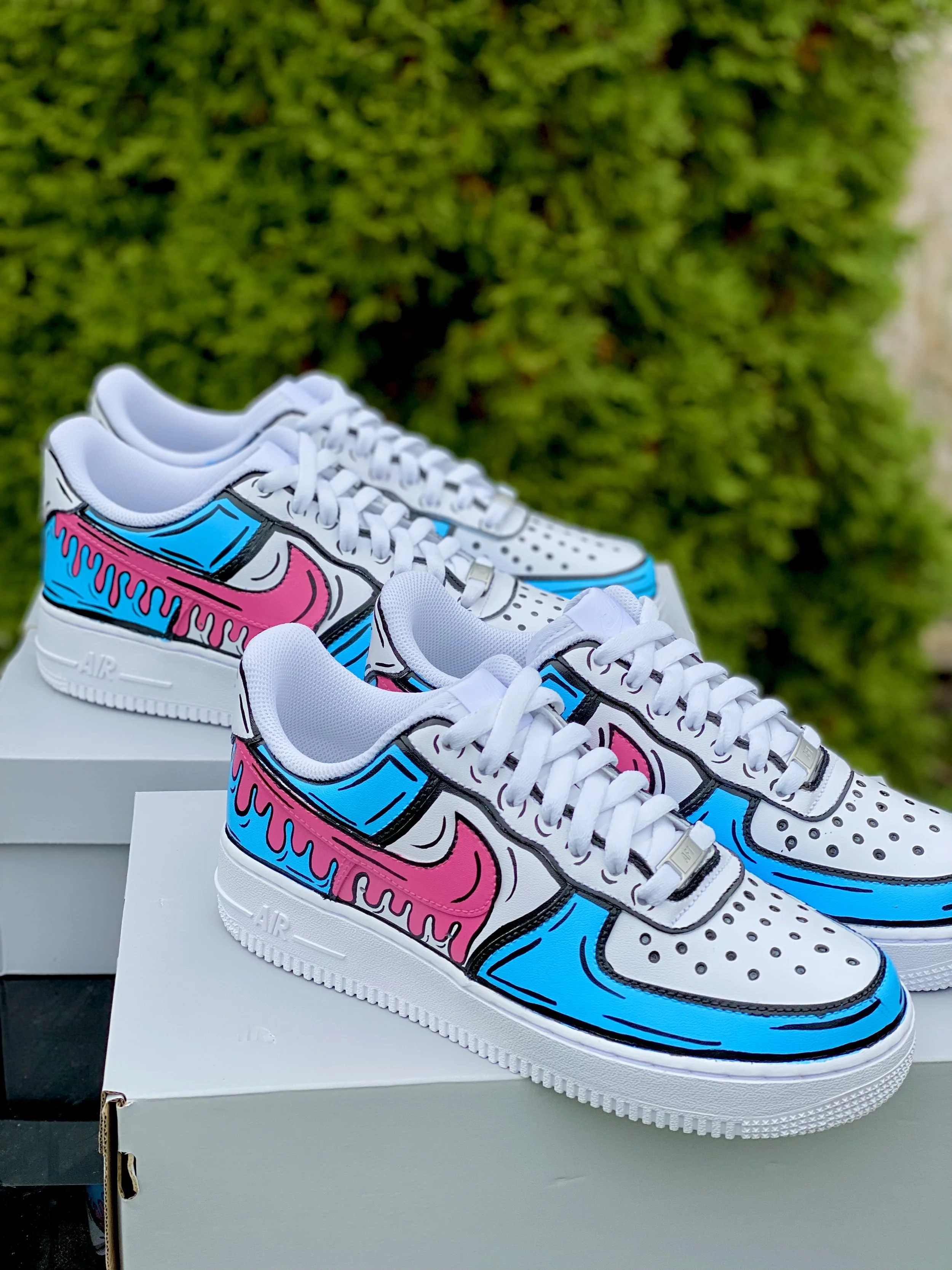 White sneakers with colorful graffiti-style design in pink and blue, placed on top of a white shoebox against a background of green bushes.