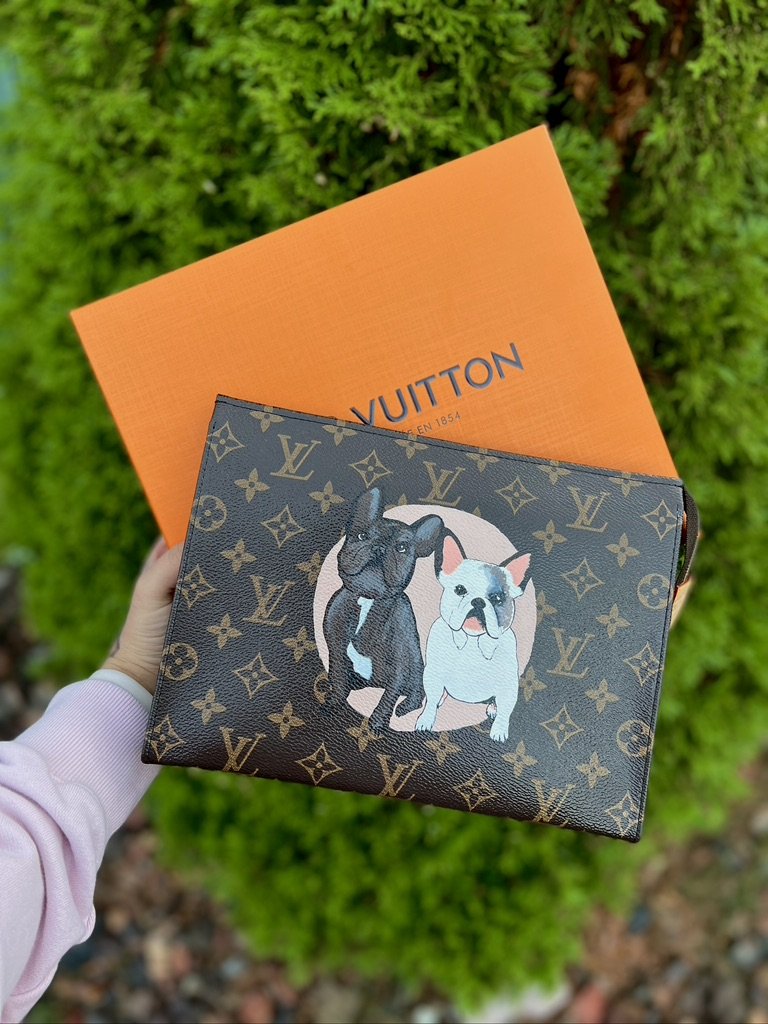 A Louis Vuitton wallet with a dog illustration on the front, held in front of an orange Louis Vuitton box, with a green, leafy background.