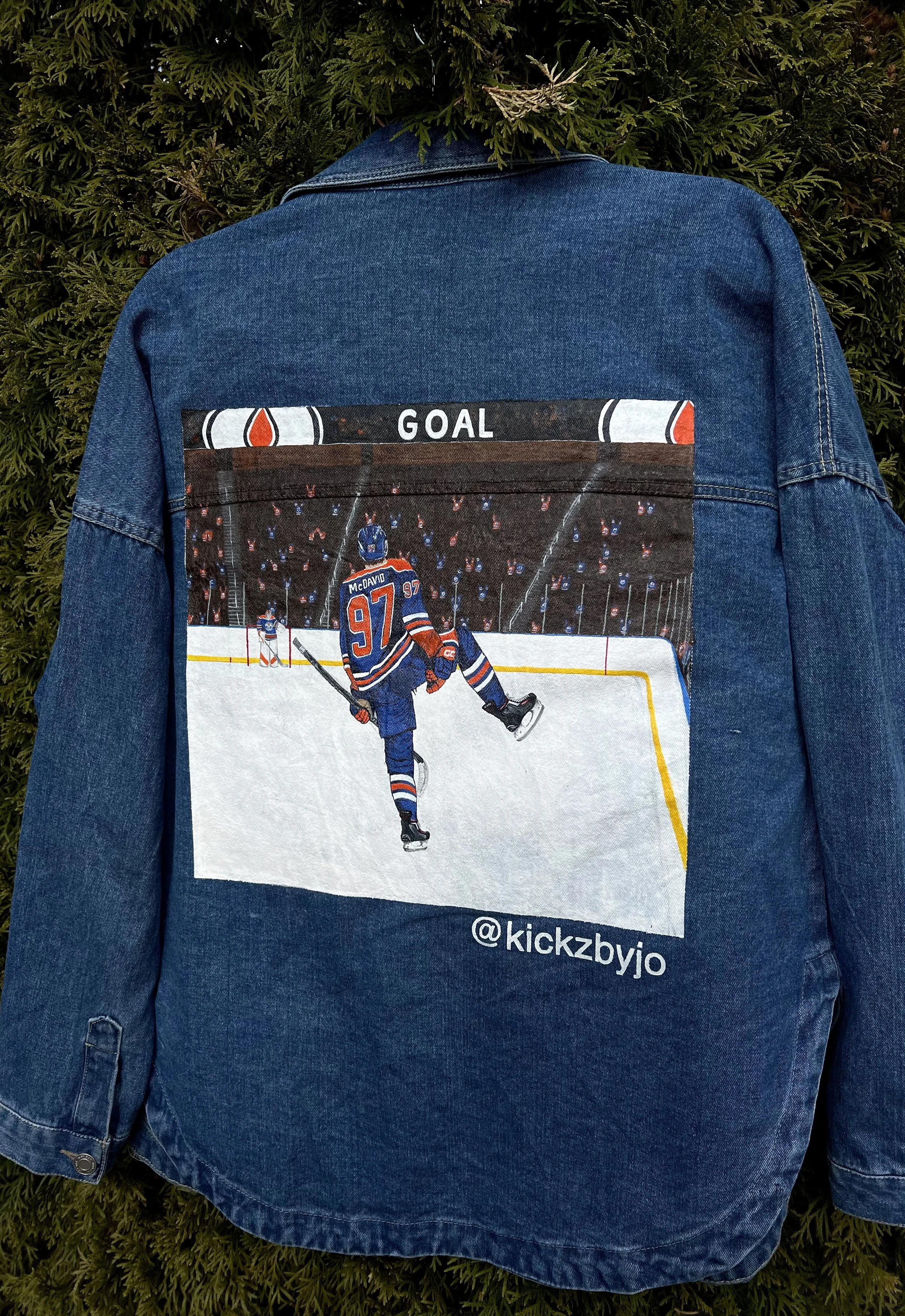 Blue denim jacket with a printed hockey game scene and the word "GOAL" on the back, hanging against green foliage.