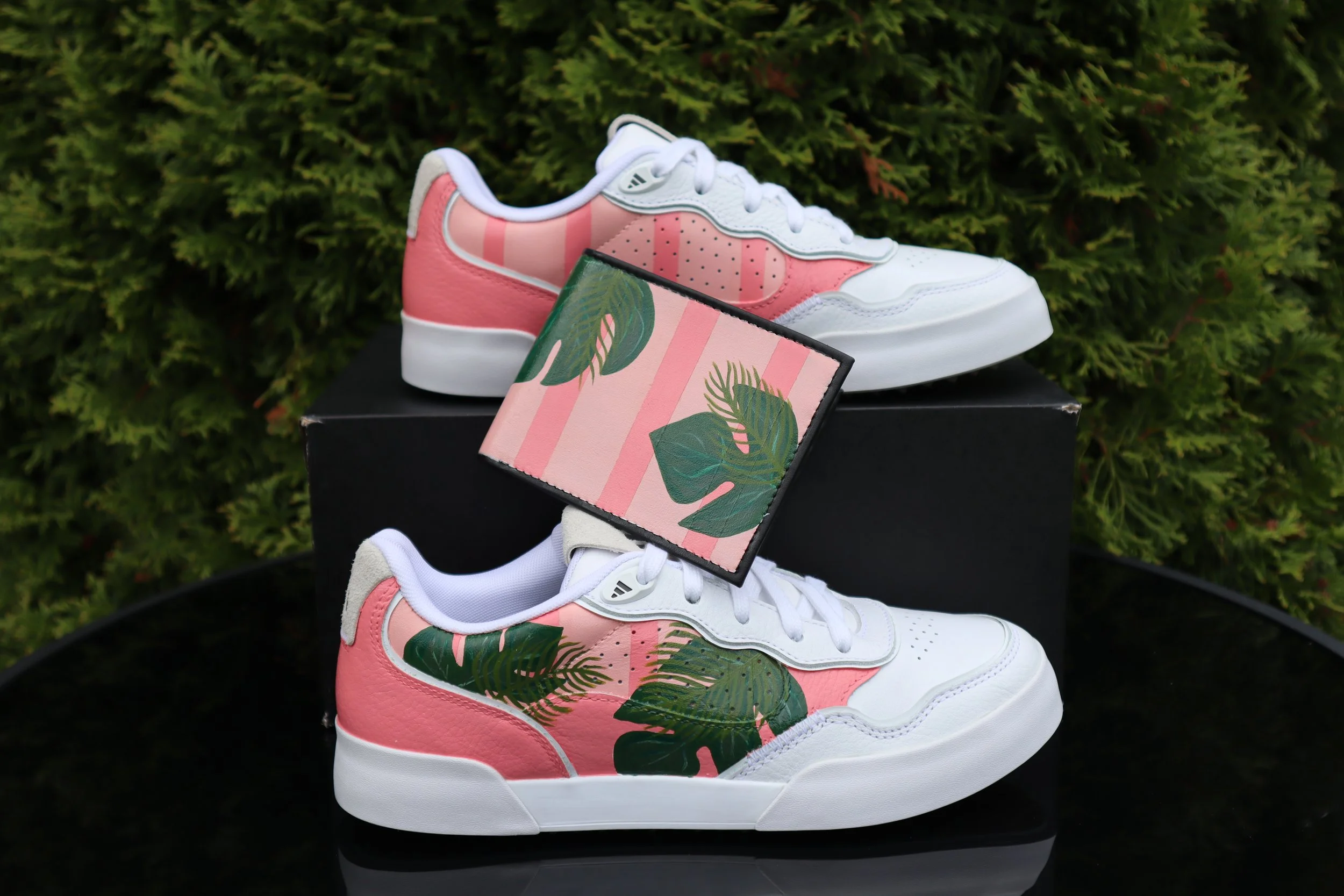 Pair of white and pink sneakers with green leaf design on the sides, placed on a black box with green foliage in the background.