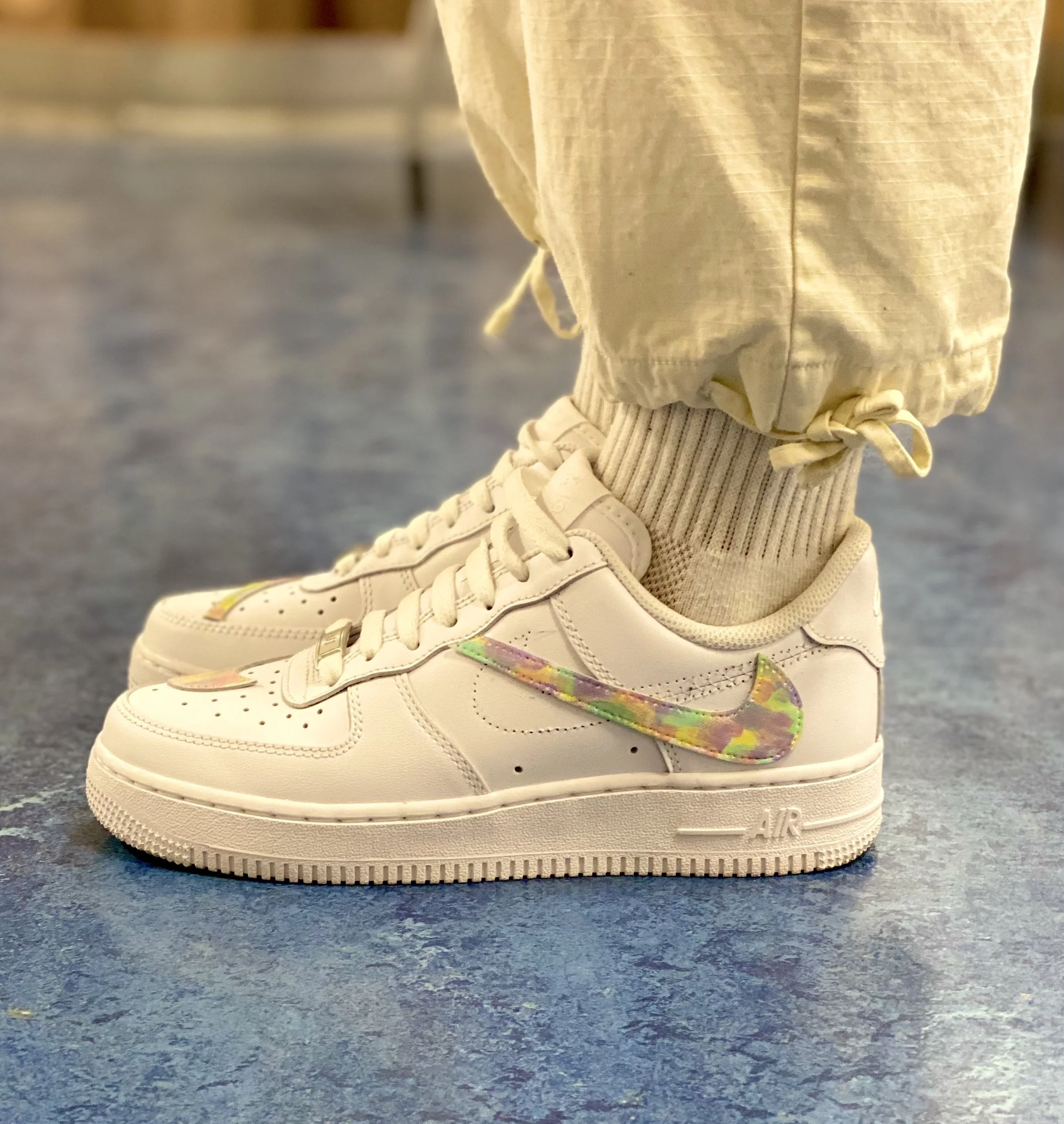 Close-up of a person's foot wearing a white sneaker with colorful details, standing on a blue speckled floor, with beige pants and a ribbed sock visible.
