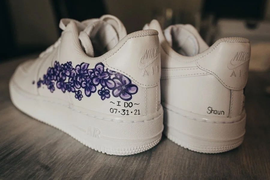 A pair of white Nike Air Force 1 sneakers with purple floral artwork and handwritten messages, including a date and a name.