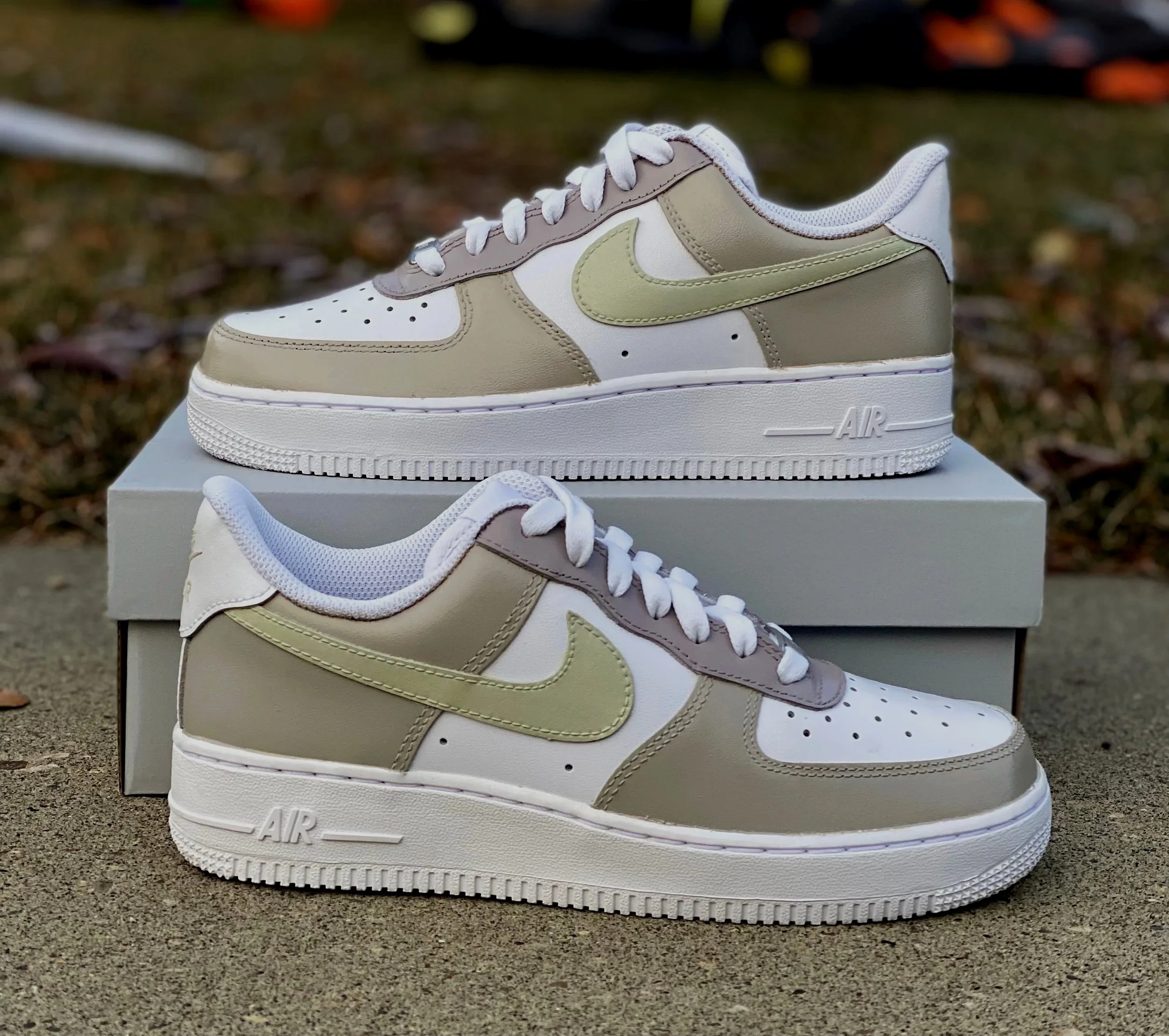 A beige and white Nike Air Force 1 sneaker with green accents on top of a shoe box outdoors on a concrete surface.