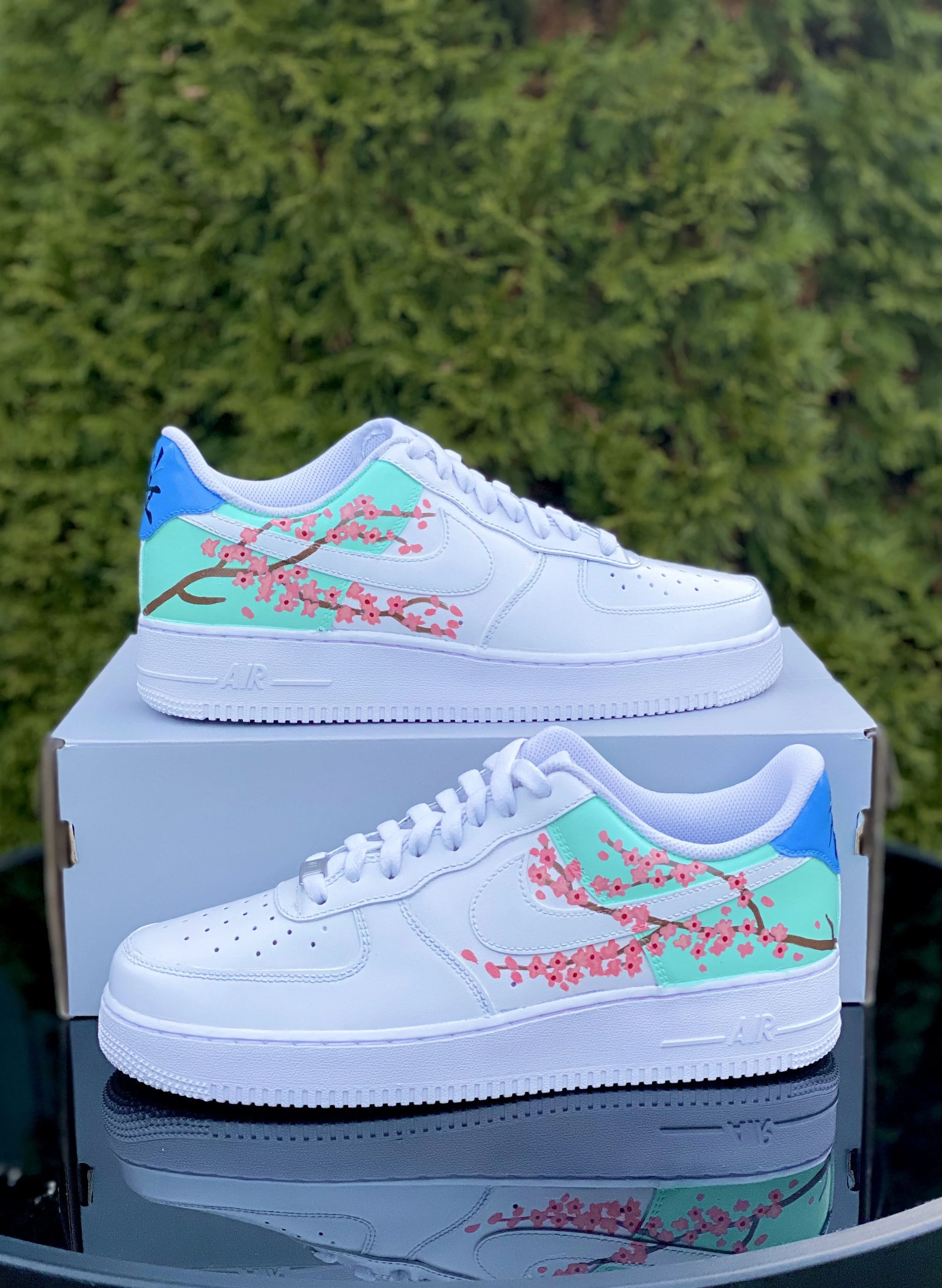 A pair of white Nike Air Force 1 sneakers with cherry blossom artwork on the sides, placed on top of a white shoebox against a blurry green background.