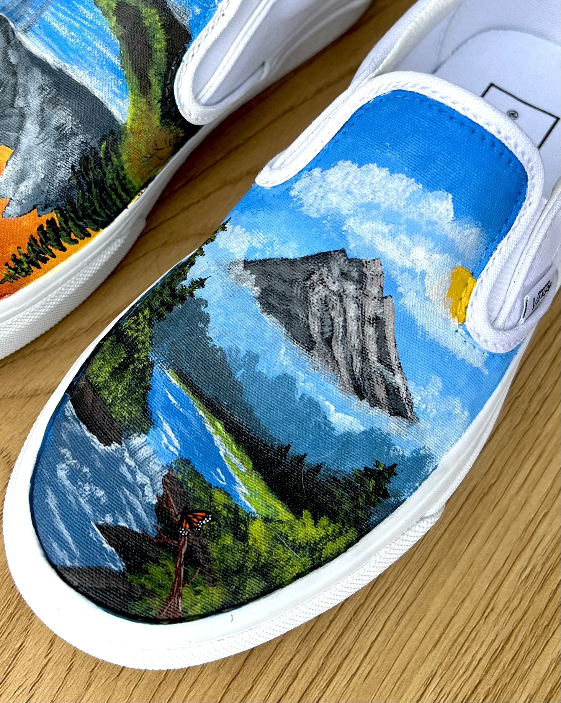 Close-up of sneakers featuring detailed landscape paintings on the canvas, depicting a mountain scene with a blue sky, clouds, and greenery.