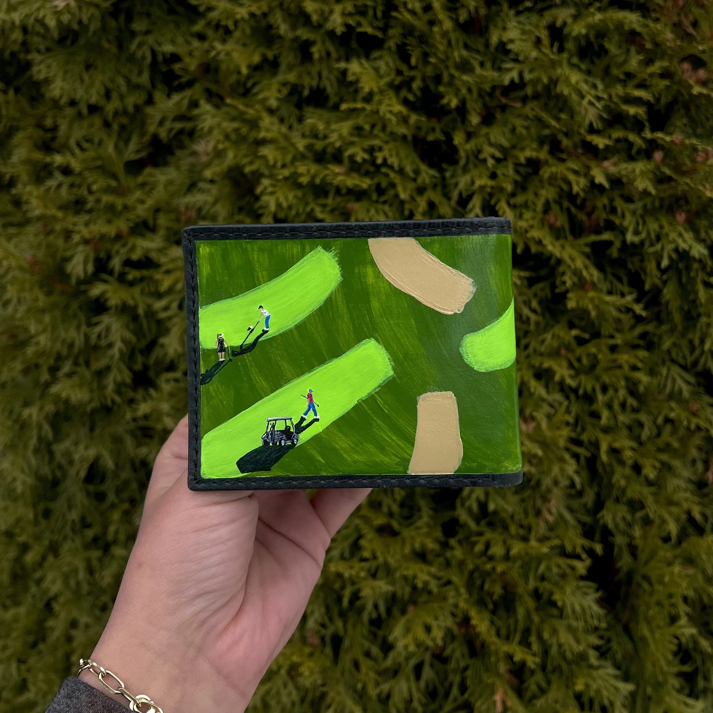A hand holding a small box with green and beige painted design, featuring illustrations of people playing golf, against a background of green foliage.