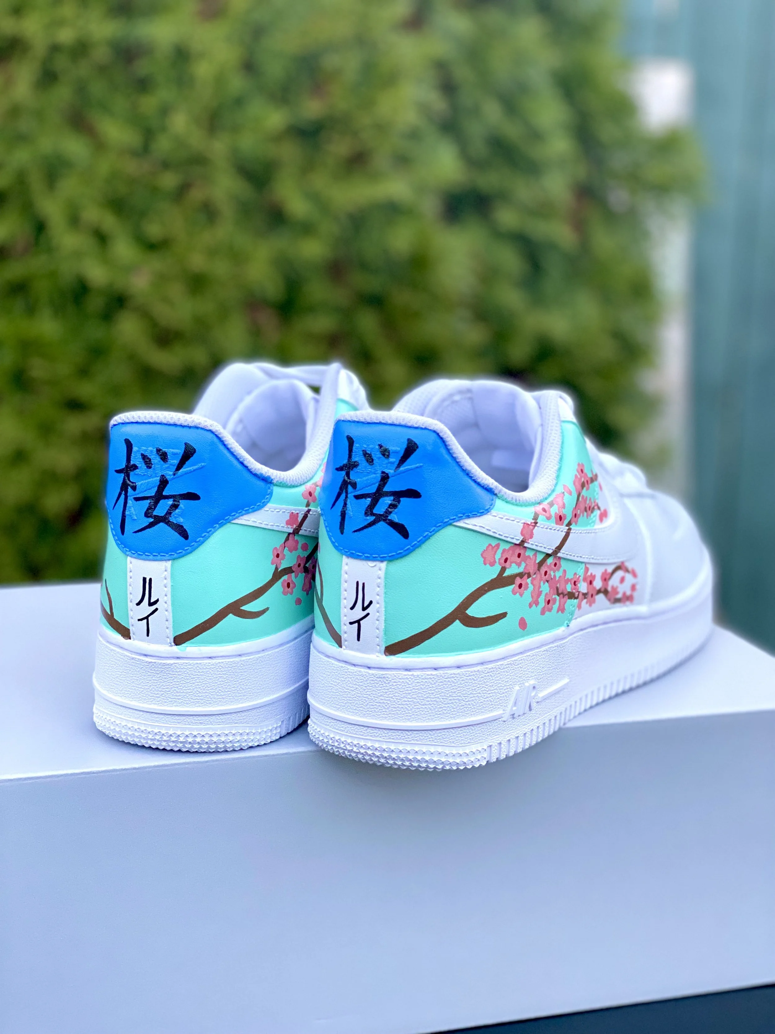 A pair of custom painted white sneakers with floral and Japanese character designs on the sides, placed outdoors on a white surface with a blurred green background.