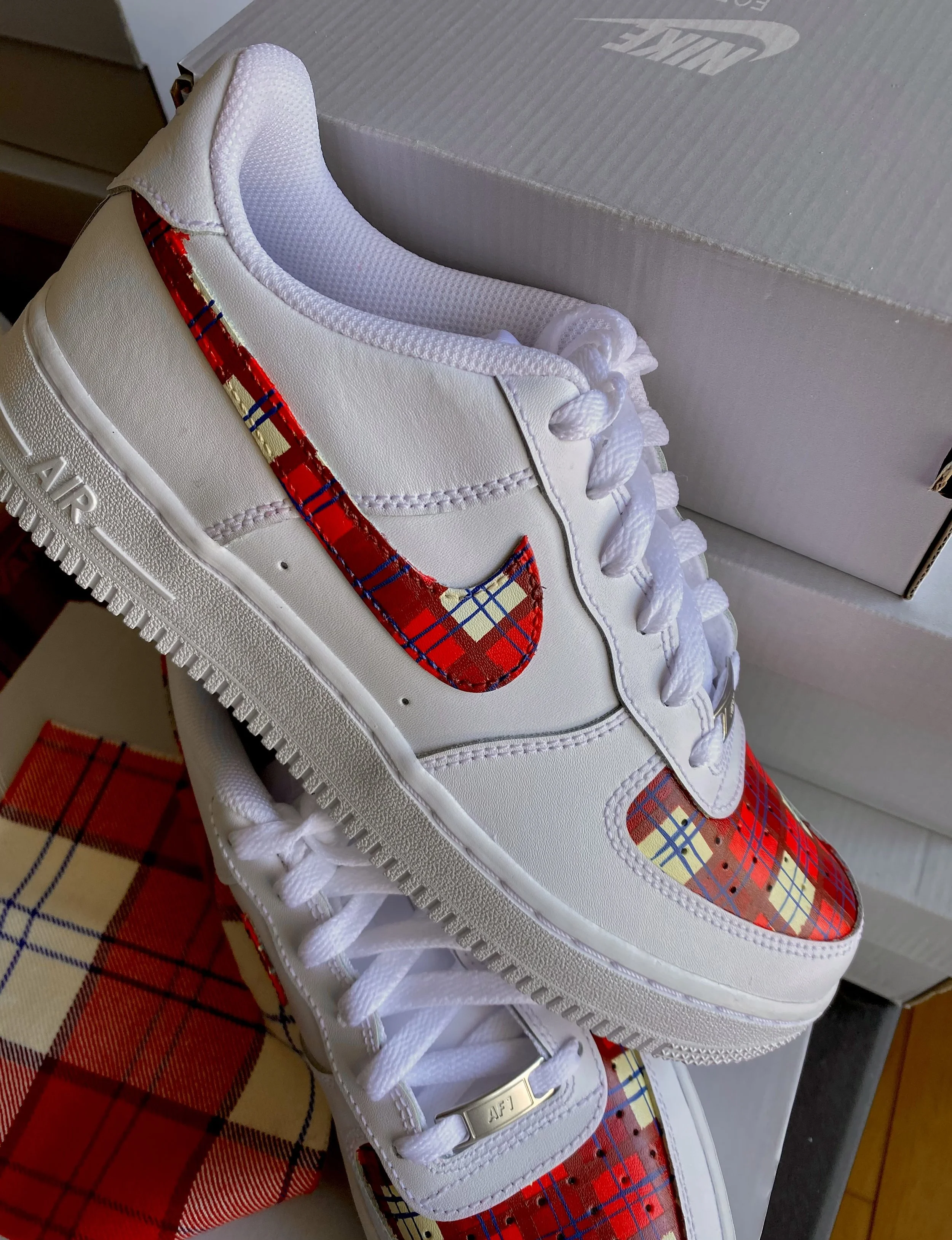 Close-up of a white Nike sneaker with a tartan pattern on the Nike swoosh logo and toe cap, placed on a surface near a Nike shoebox.
