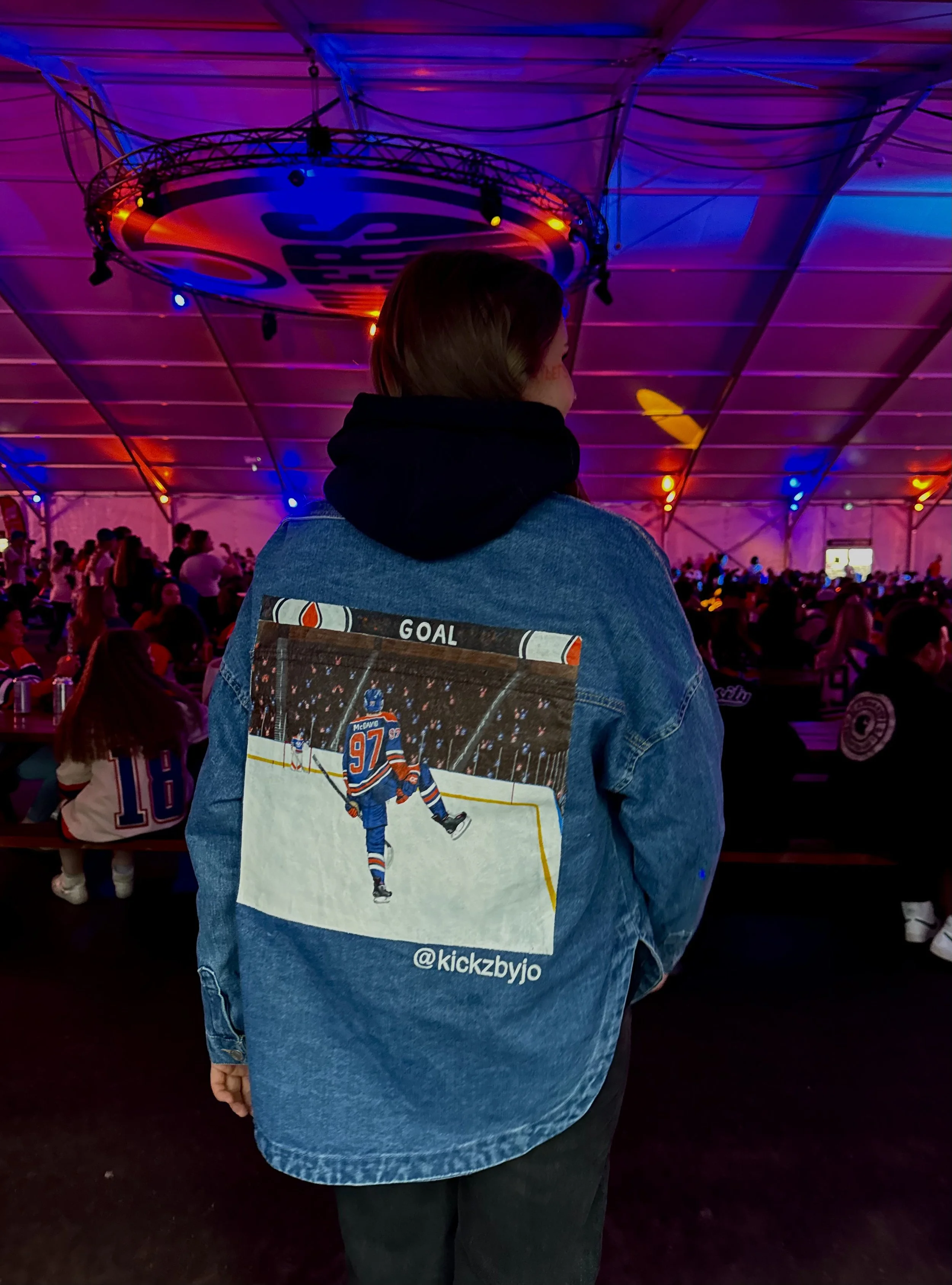 Back of a person wearing a denim jacket with a hockey player graphic on the back, standing in a large indoor venue decorated with colorful lights, with a crowd seated behind.