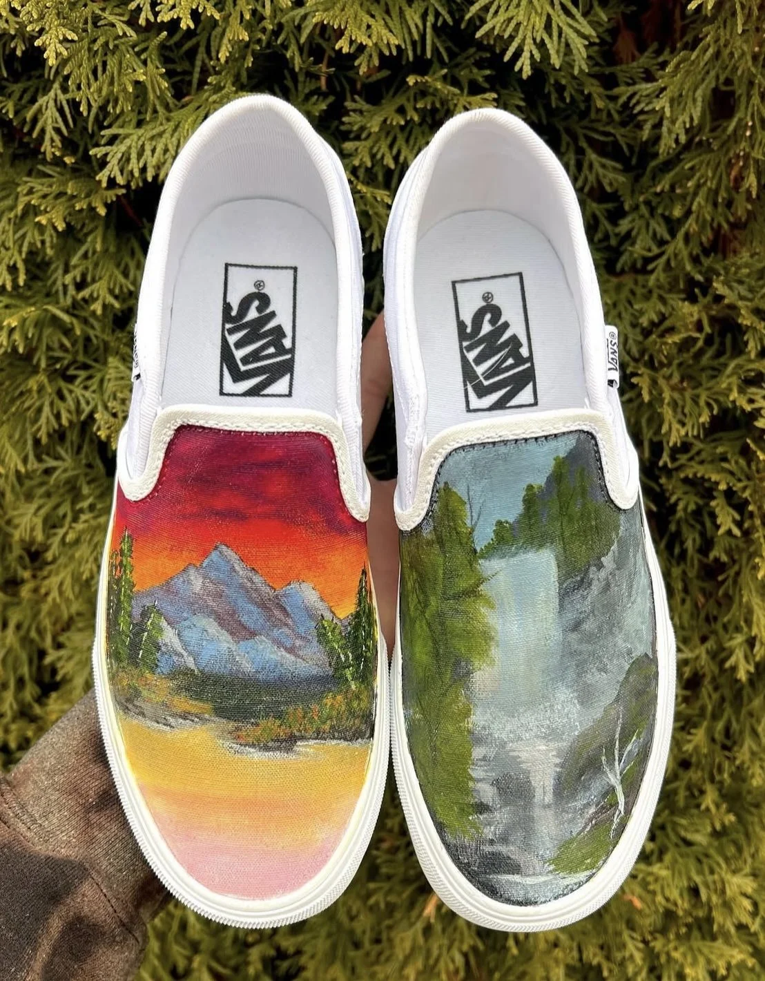 Pair of white Vans sneakers with landscape painting on the toes; the left shoe depicts mountains and a colorful sunset, the right shoe features a forest and waterfall scene.