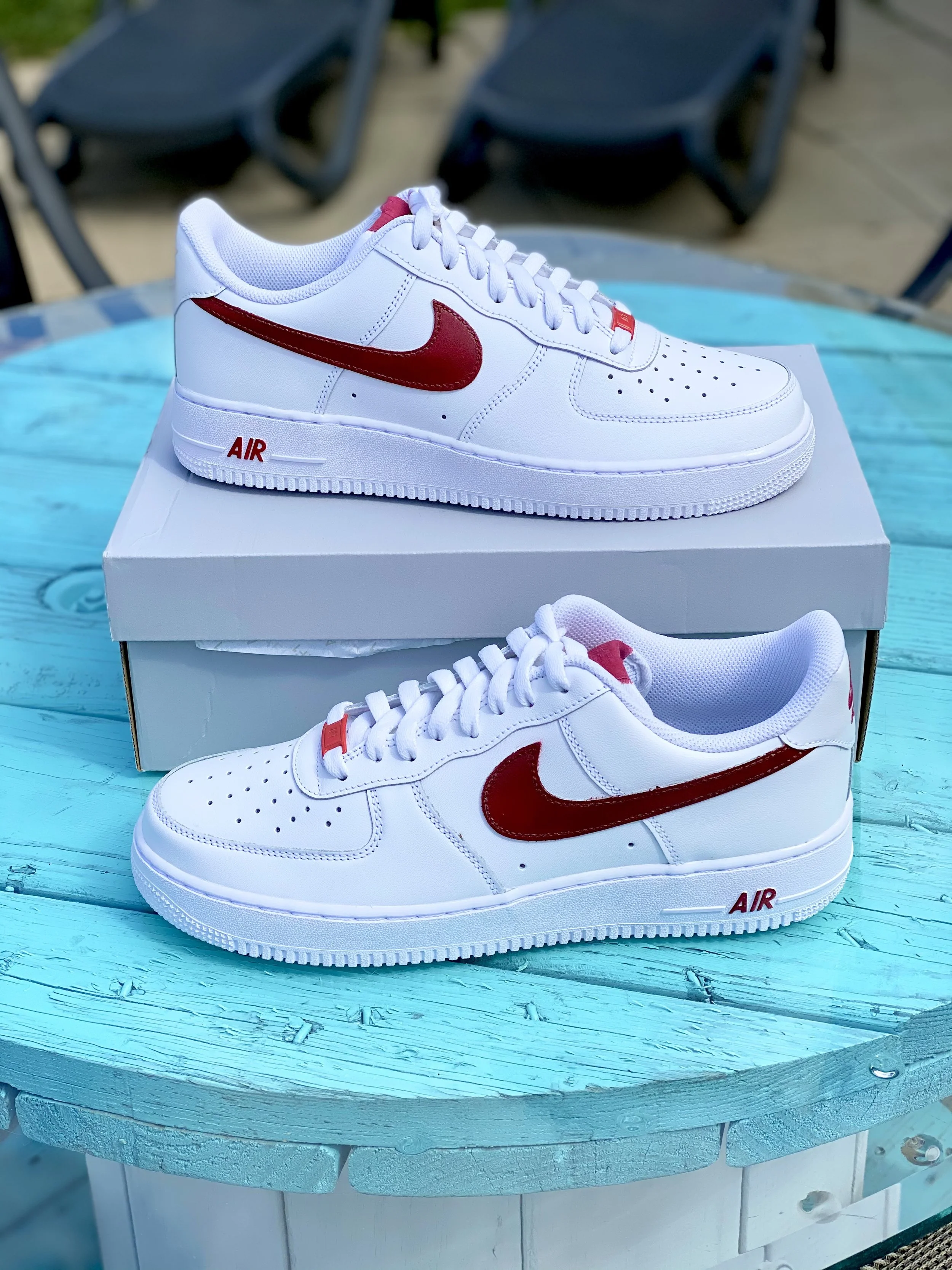 A pair of white Nike sneakers with red accents, sitting on a white shoe box on a turquoise wooden table, with black chairs in the background.