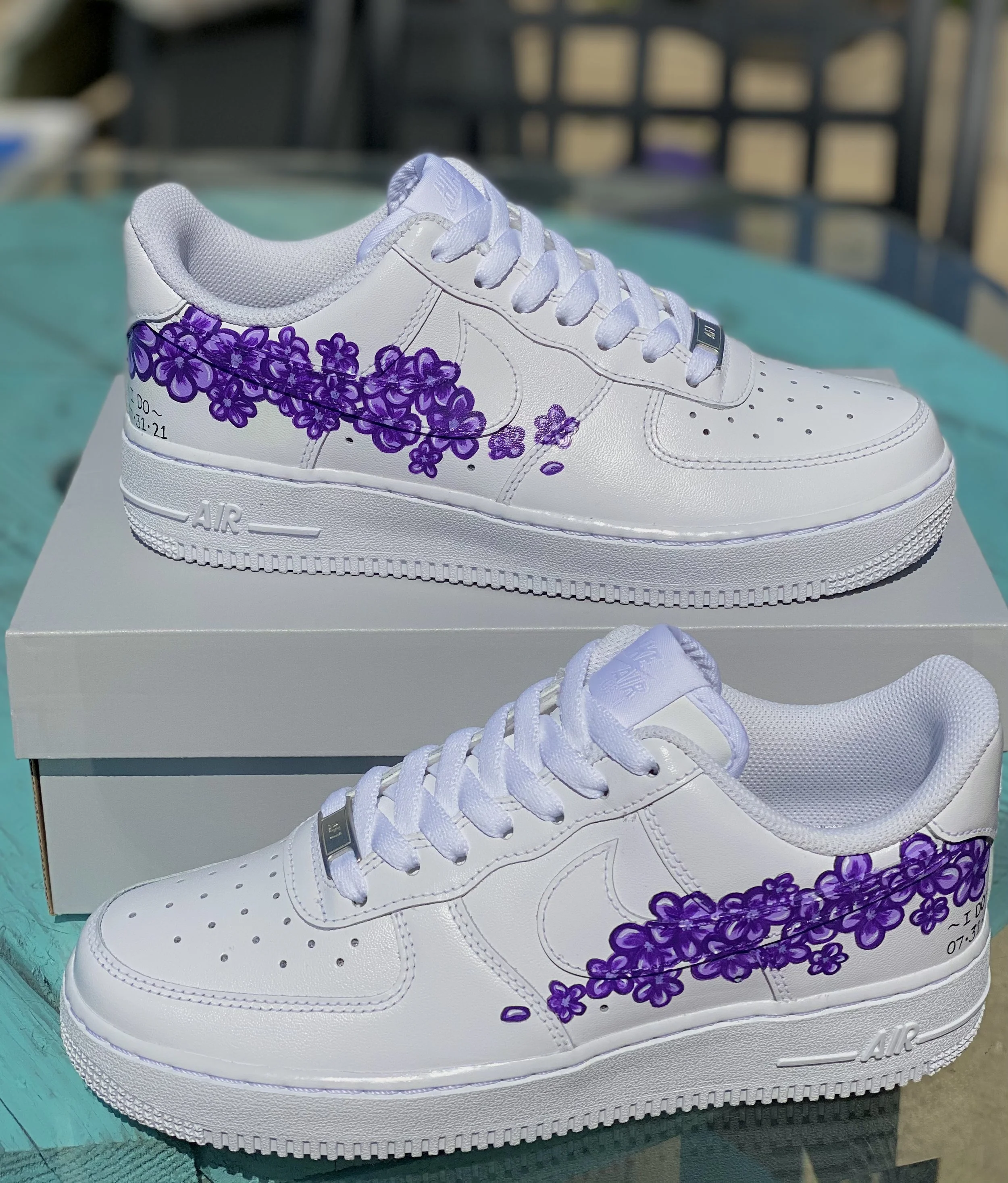 White Nike Air Force 1 sneakers with purple floral designs on the side and handwritten message, resting on a white box.