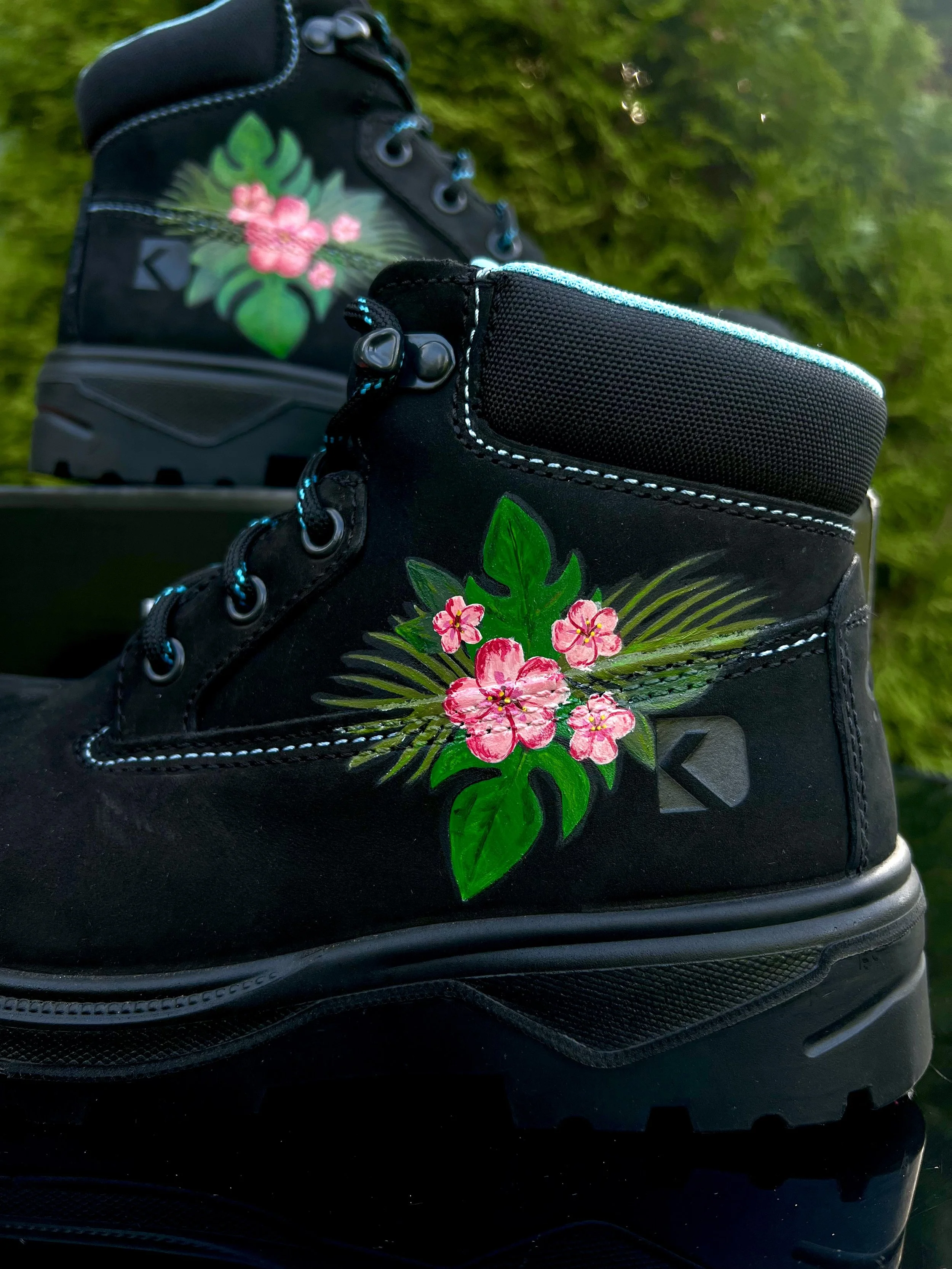 Close-up of a black hiking boot with a painted pink hibiscus flower and green leaves on the side. The background is blurred green foliage.
