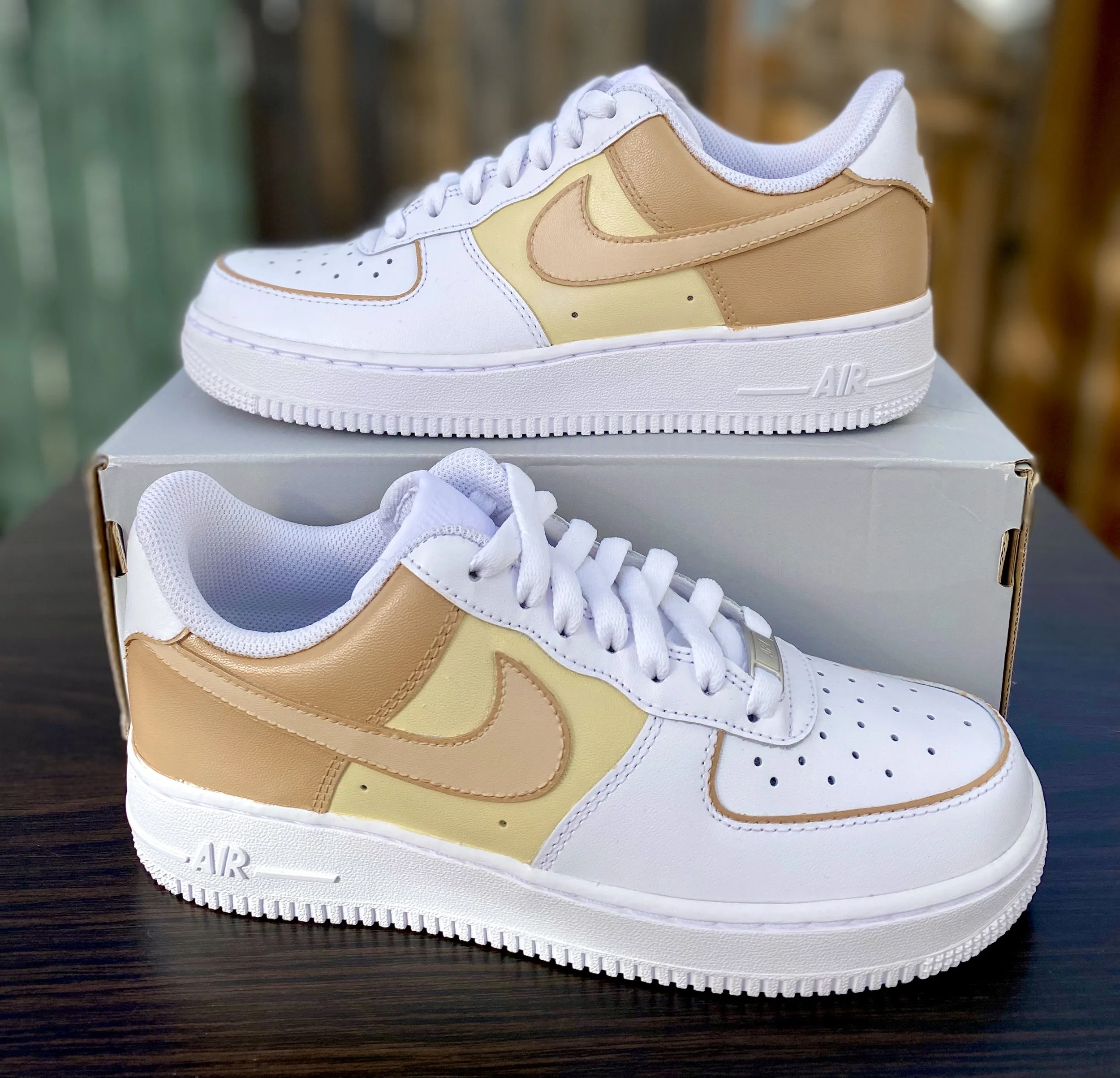 Close-up of a white and tan Nike Air Force 1 sneaker on a dark wooden surface, with a shoebox in the background.