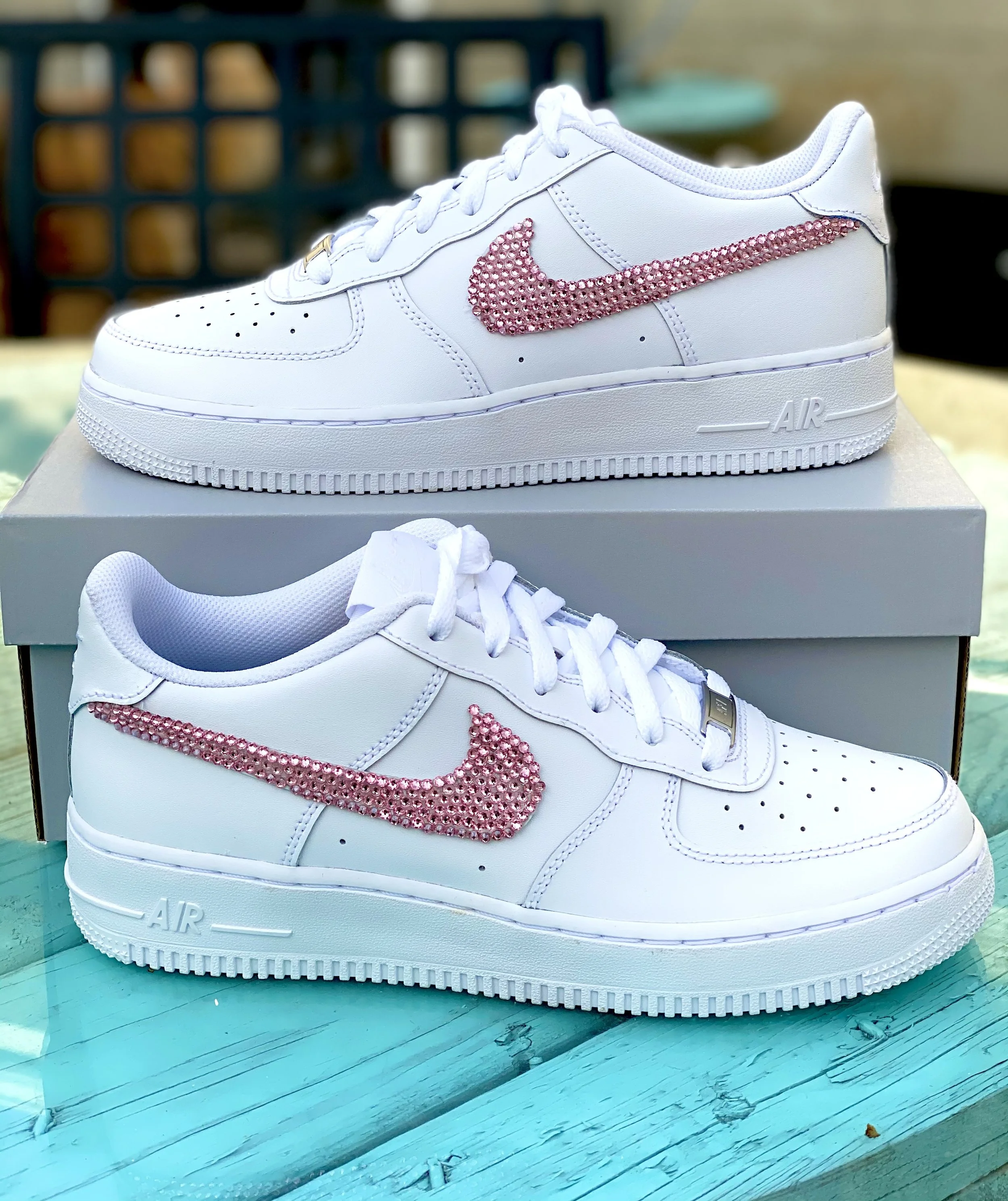 White Nike Air Force 1 sneakers decorated with pink rhinestones to form the Nike swoosh logo, resting on a shoebox on a blue wooden surface.