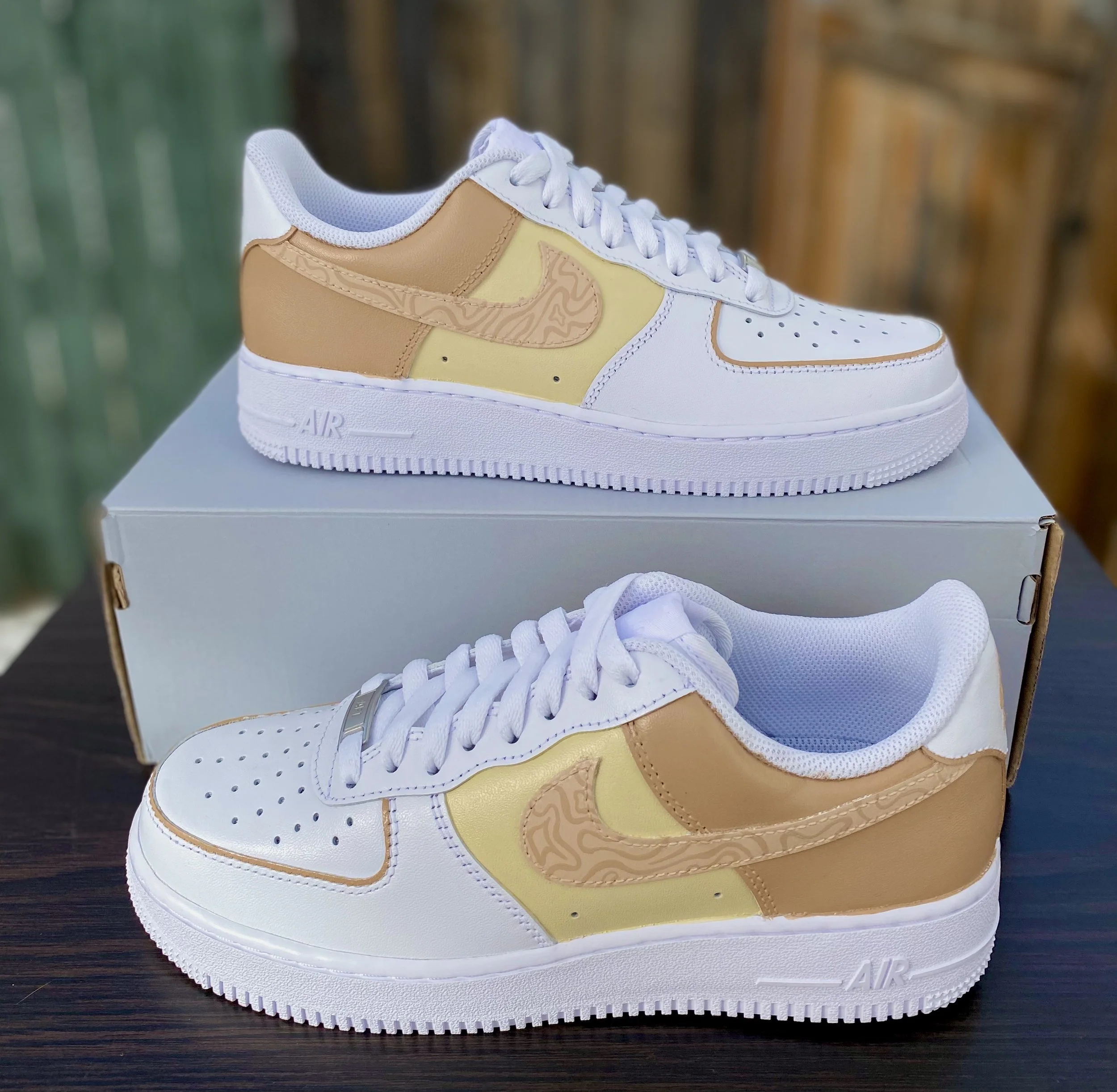 A white Nike Air Force 1 sneaker with beige and light yellow accents, placed on top of a white Nike shoebox, with a blurred background of wooden and green surfaces.