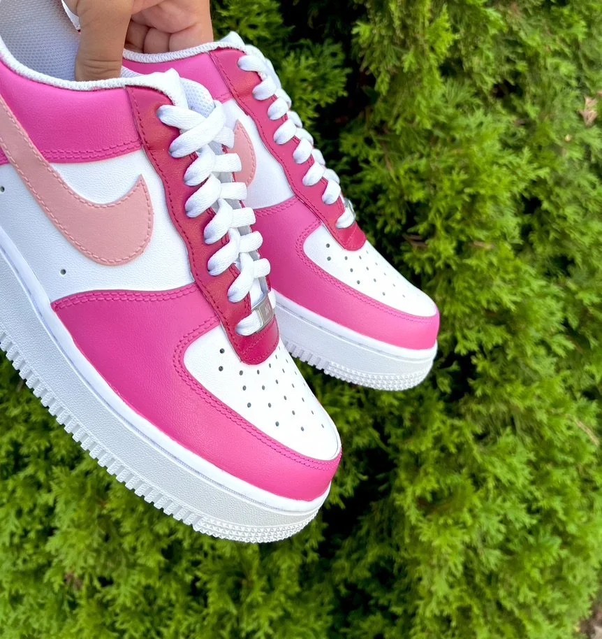 Close-up of a pair of pink and white Nike sneakers held by a person against a green bush background.