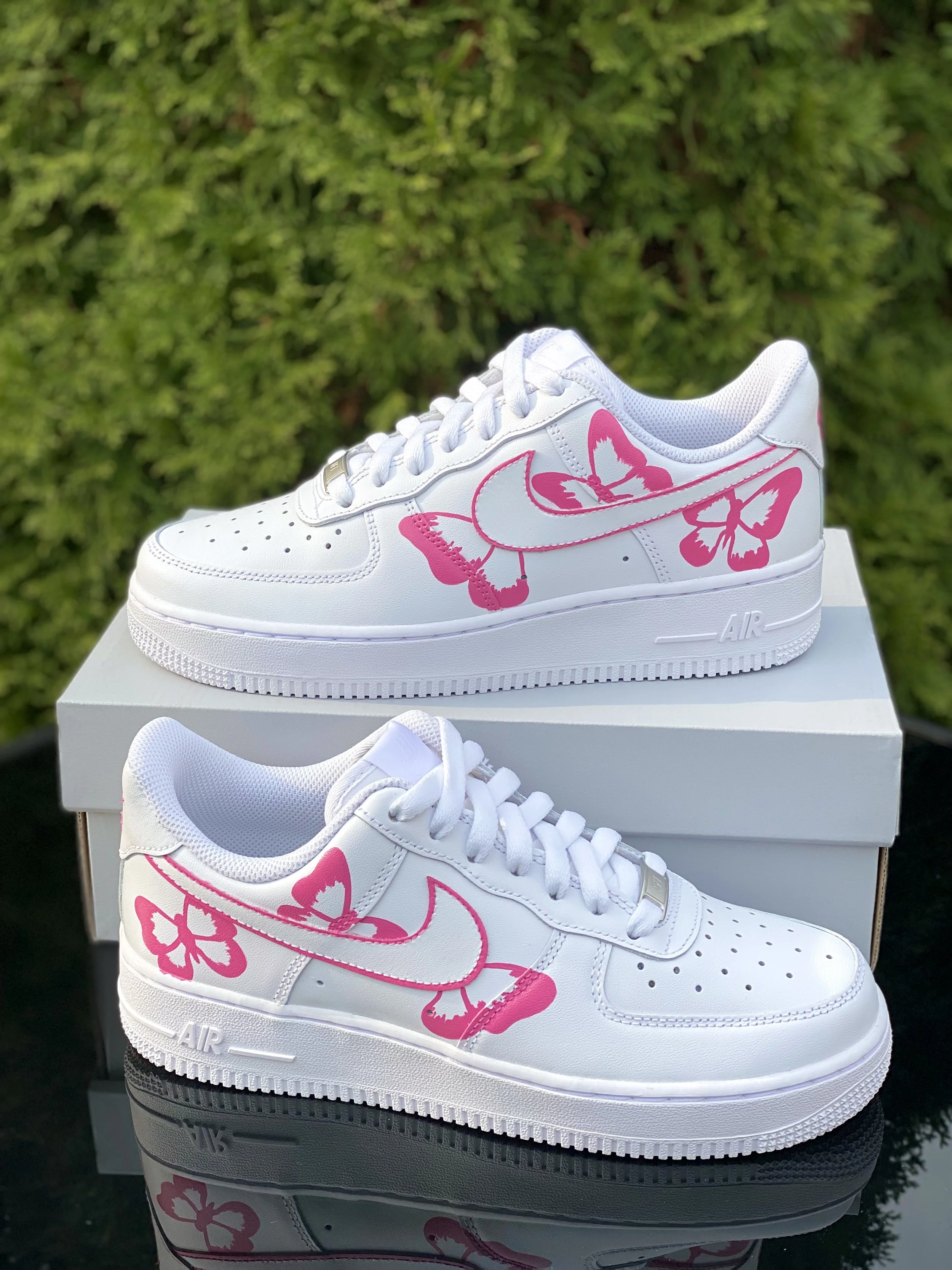 A pair of white sneakers with pink butterfly designs on the sides, placed on top of a white shoebox with a reflection on a black surface, against a green leafy background.