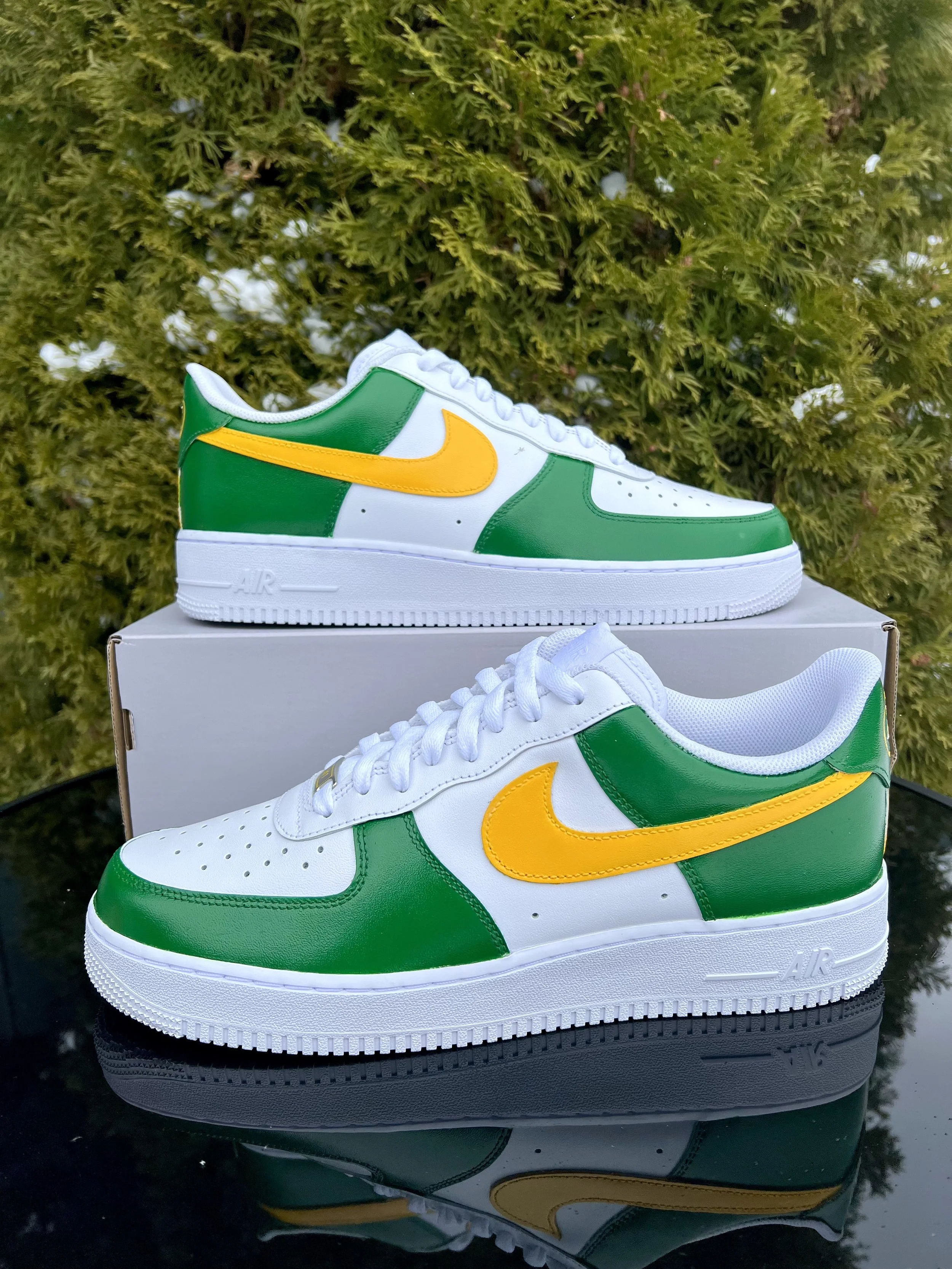 Green and white sneakers with yellow Nike swoosh, placed on top of a white shoebox, with a bush in the background and a reflection of the shoes on a shiny surface.