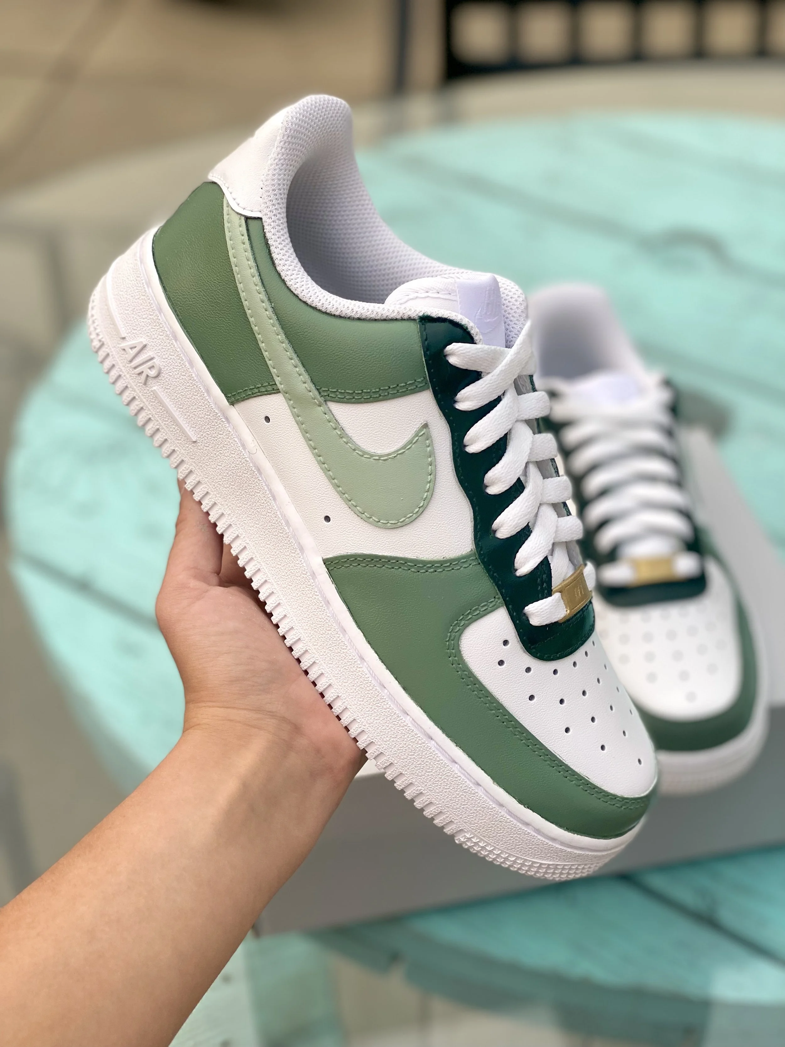 Close-up of a white and green sneaker with white laces, held by a hand, with another similar sneaker in the background.