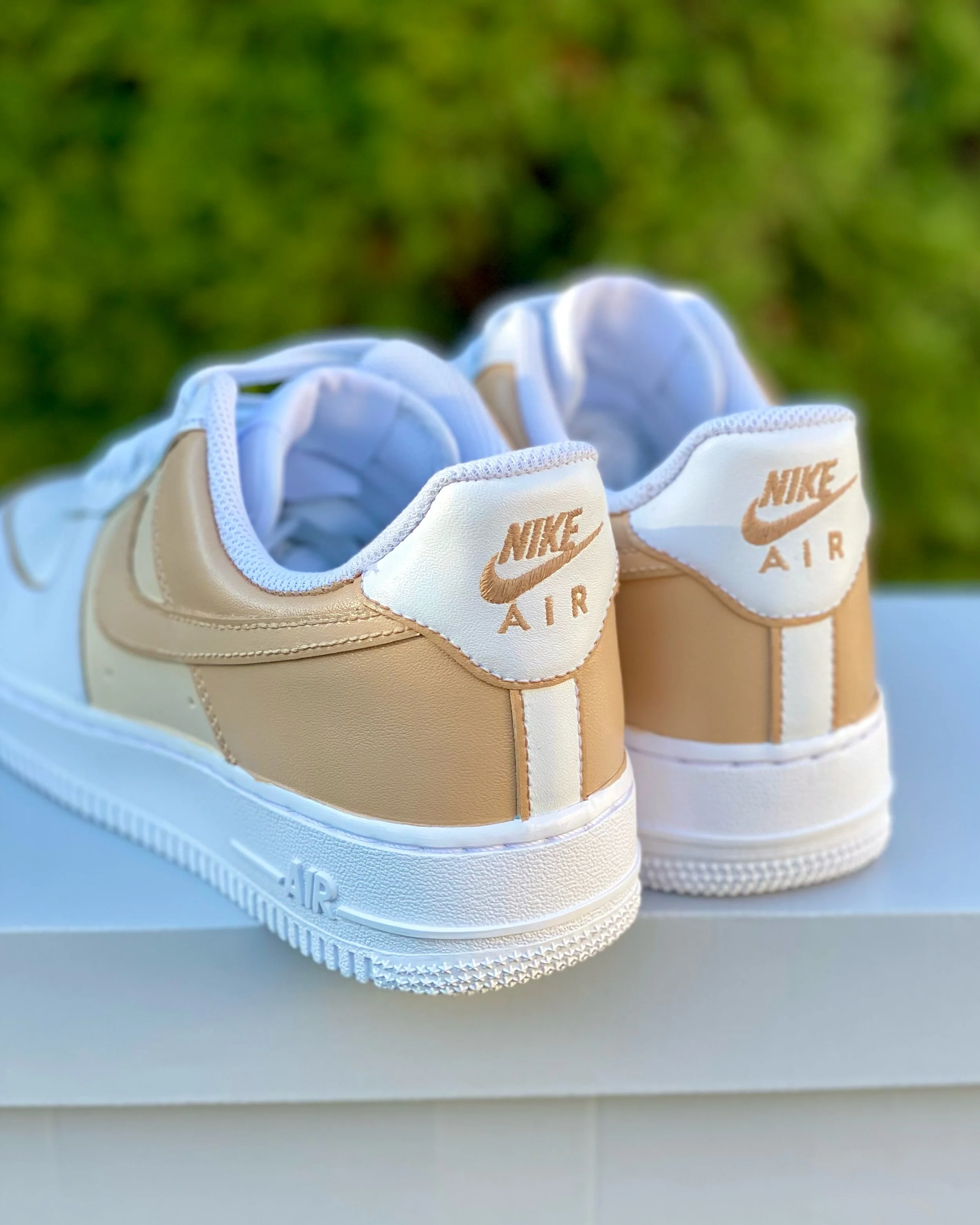 Close-up of beige and white Nike Air sneakers with light brown accents, placed on a gray surface with a blurred green background.